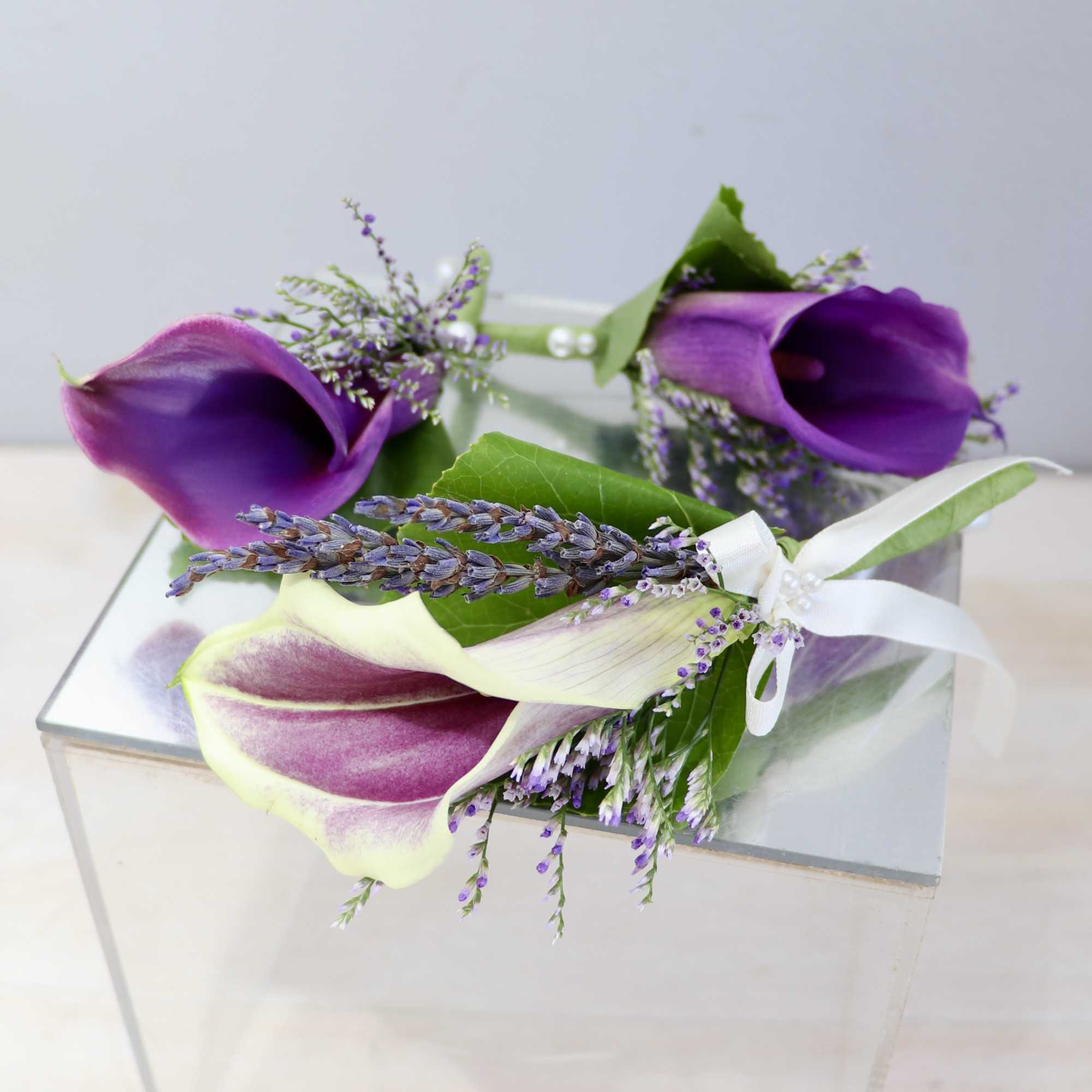 Set of purple and cream mini calla lily boutonnieres with lavender accents and ribbon on a mirrored glass cube