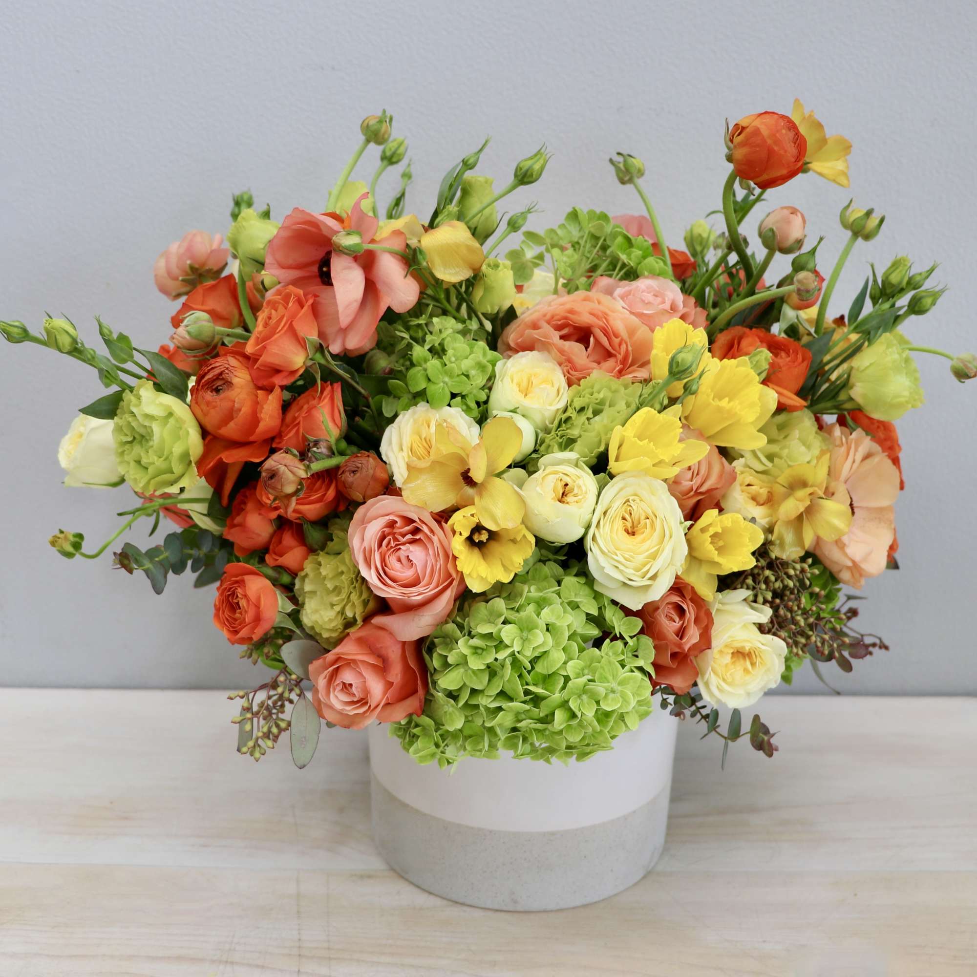 Low round arrangement of peach, yellow, orange, and green flowers in a white ceramic vase