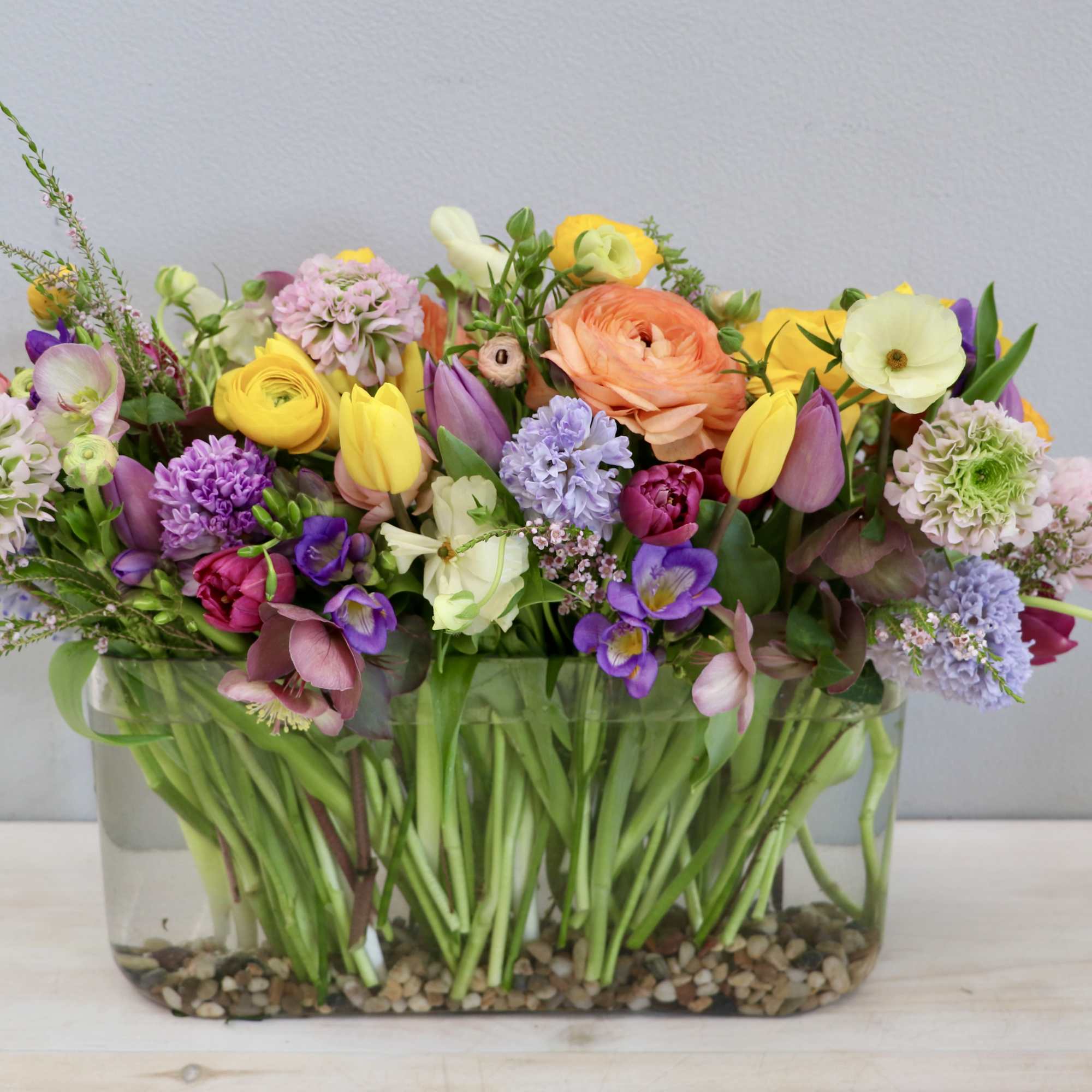Low rectangular glass vase filled with assorted colorful spring flowers in yellow, pink, purple, and orange.