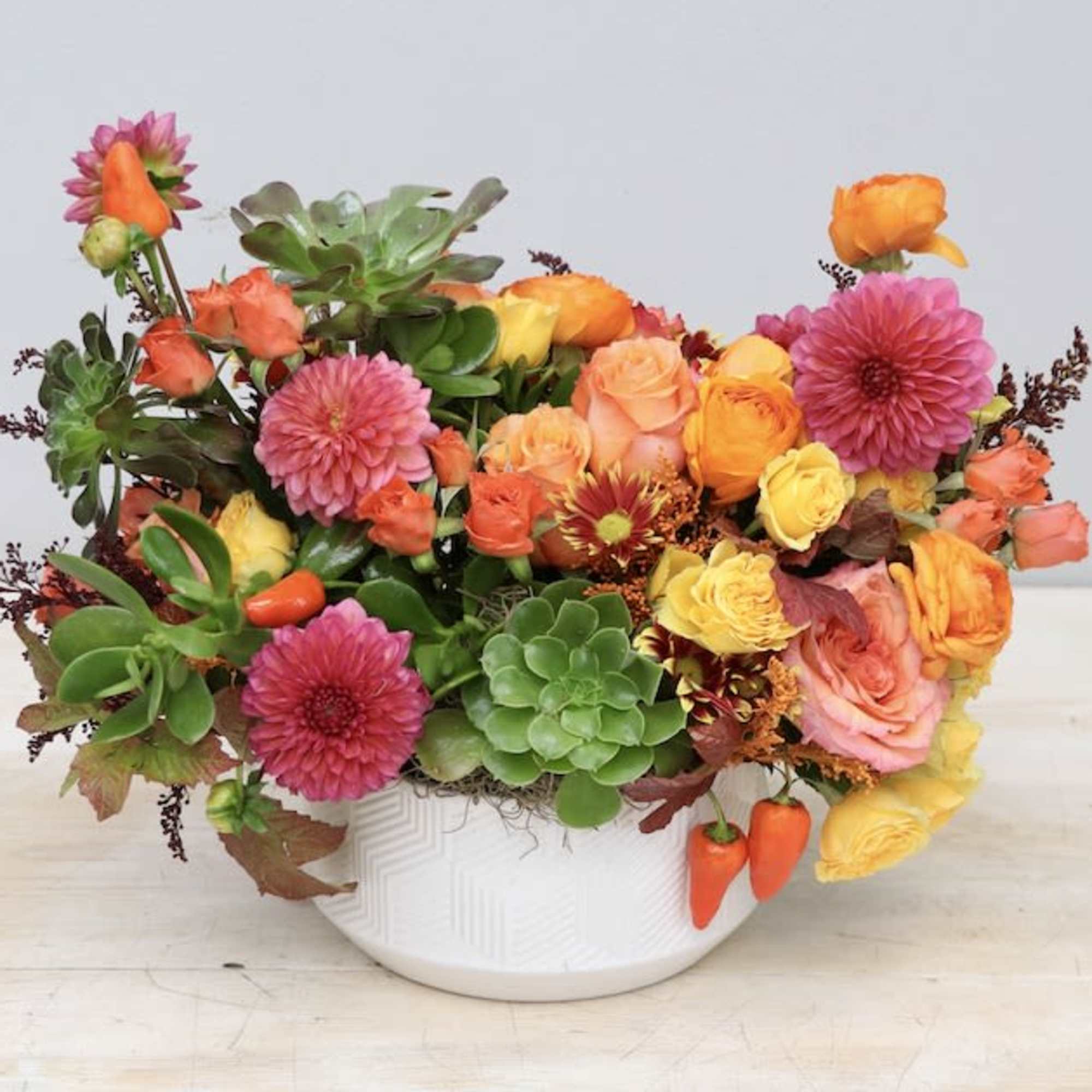 Low arrangement of orange and yellow roses, pink dahlias, and green succulents in a white bowl vase