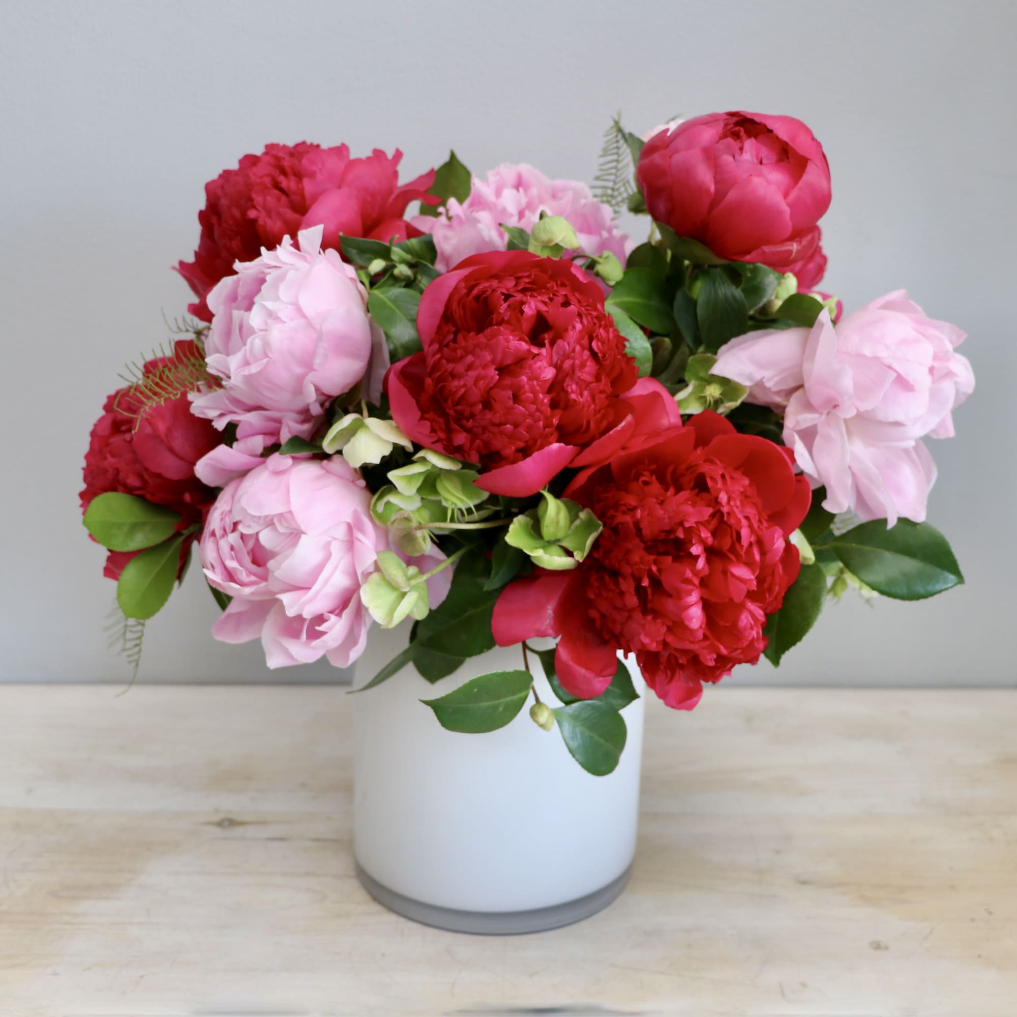 Red and pink peony arrangement in a white cylinder vase on a light wood surface