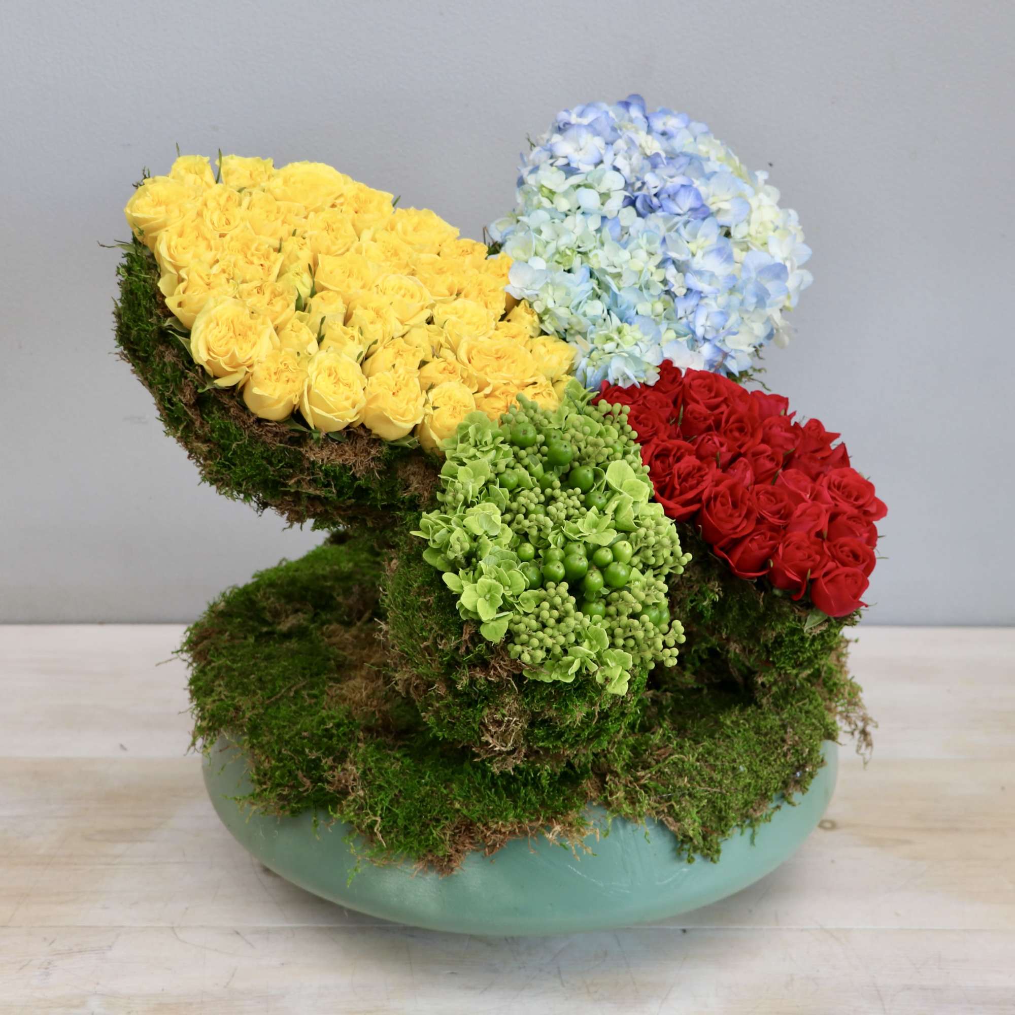 Modern arrangement with yellow and red roses and blue hydrangeas on moss in a low green bowl