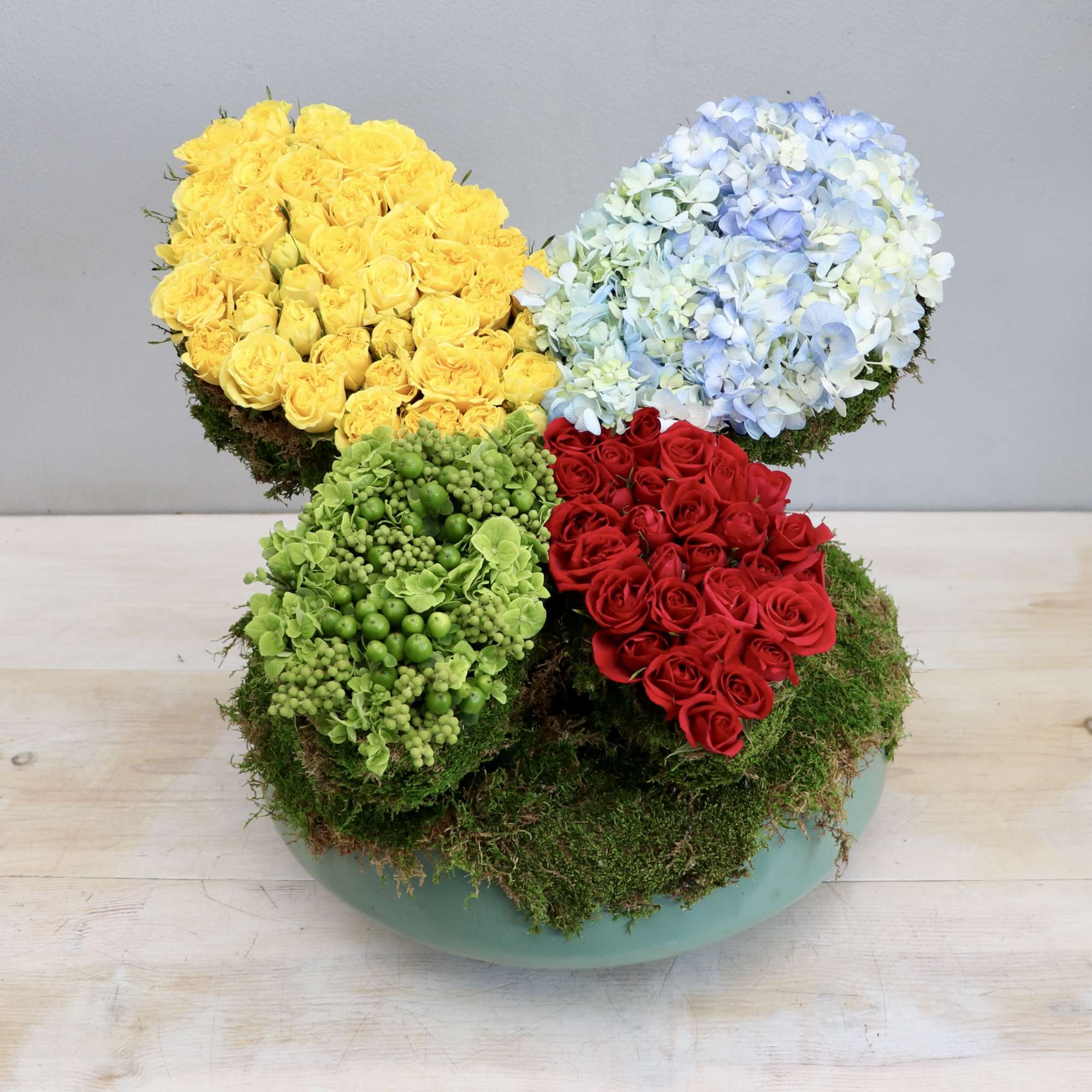 Modern low arrangement with yellow and red roses, blue hydrangeas, and green berries in a round bowl