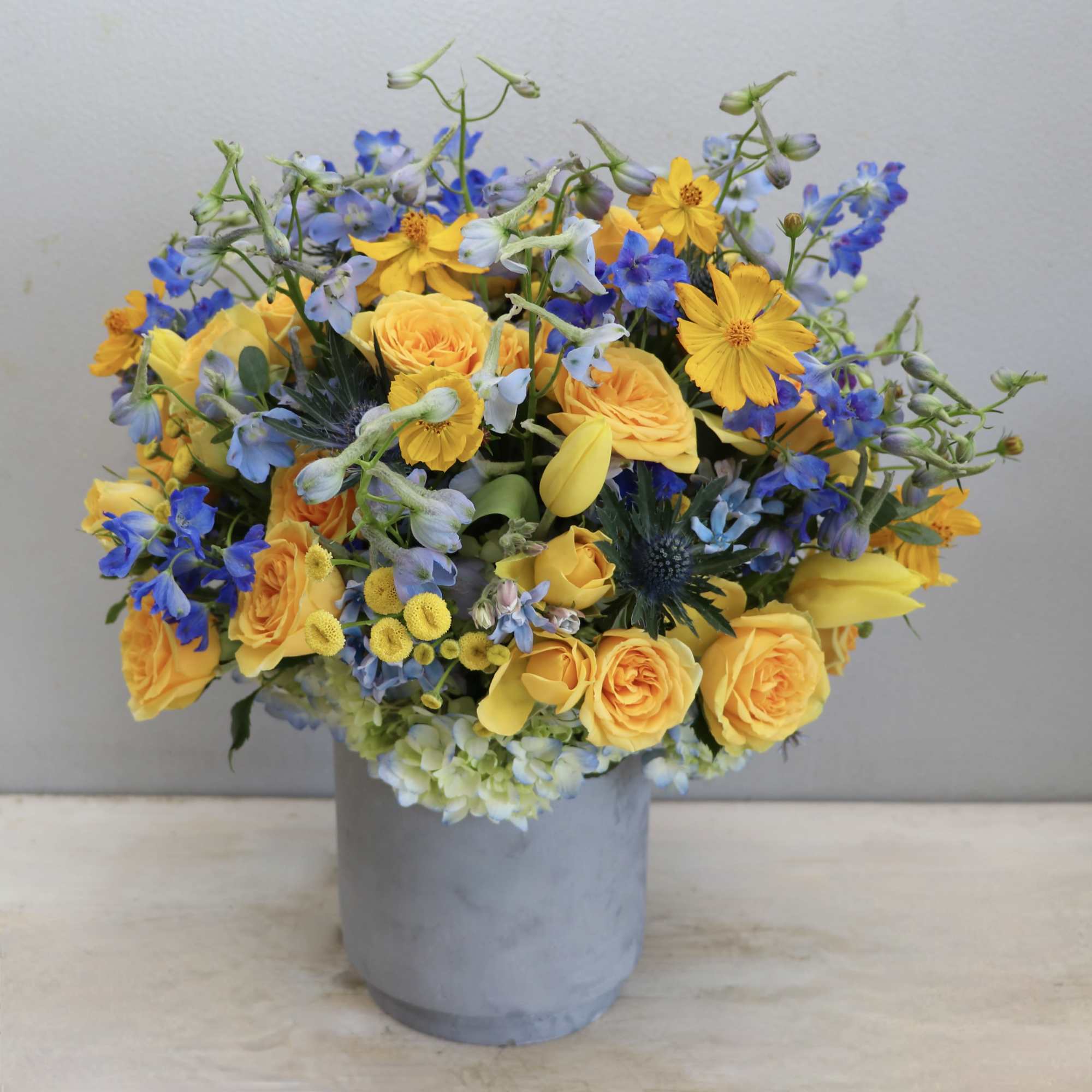 Yellow and blue mixed flower arrangement in a gray cylinder vase