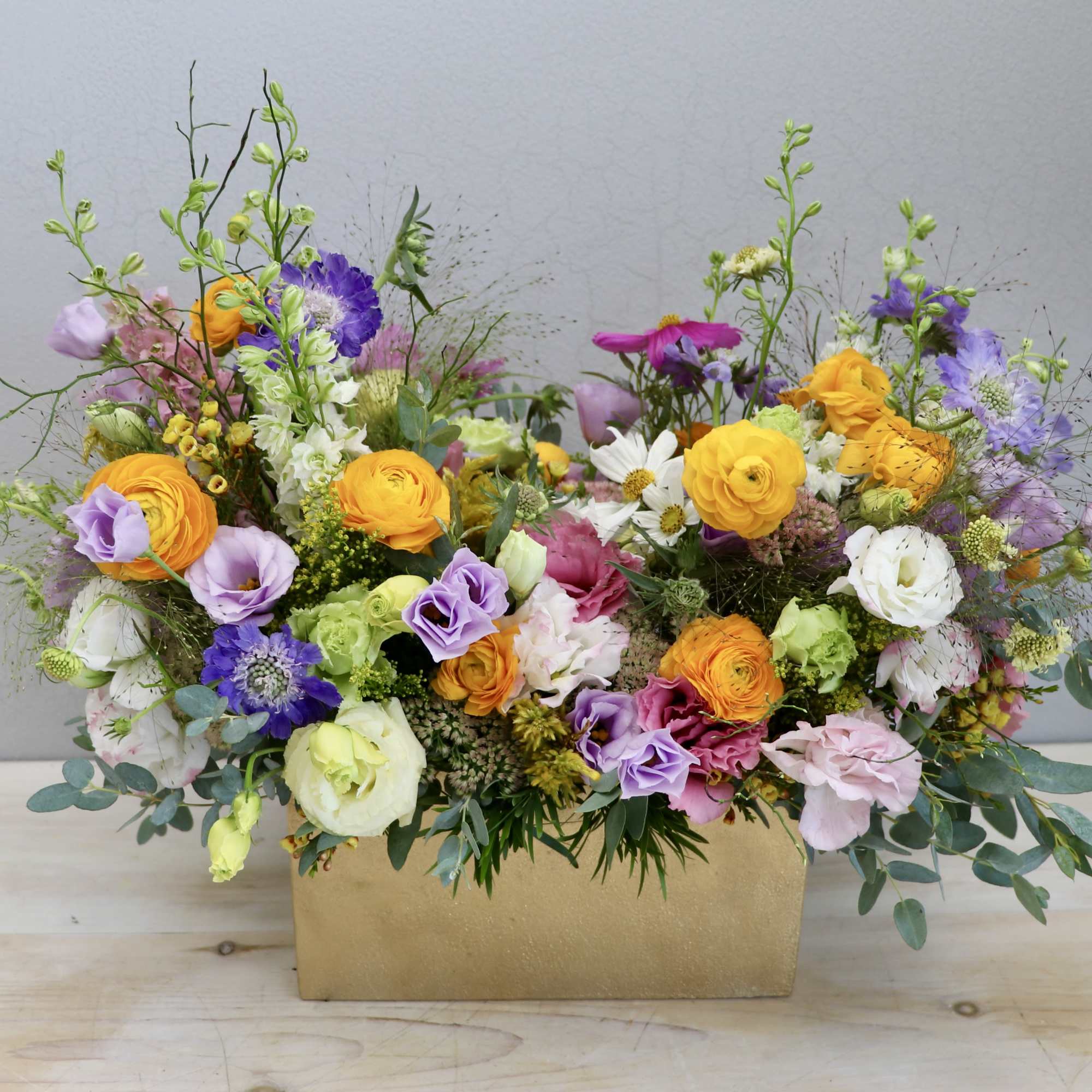 Colorful mixed flower arrangement with yellow, pink, and purple blooms in a rectangular gold container.