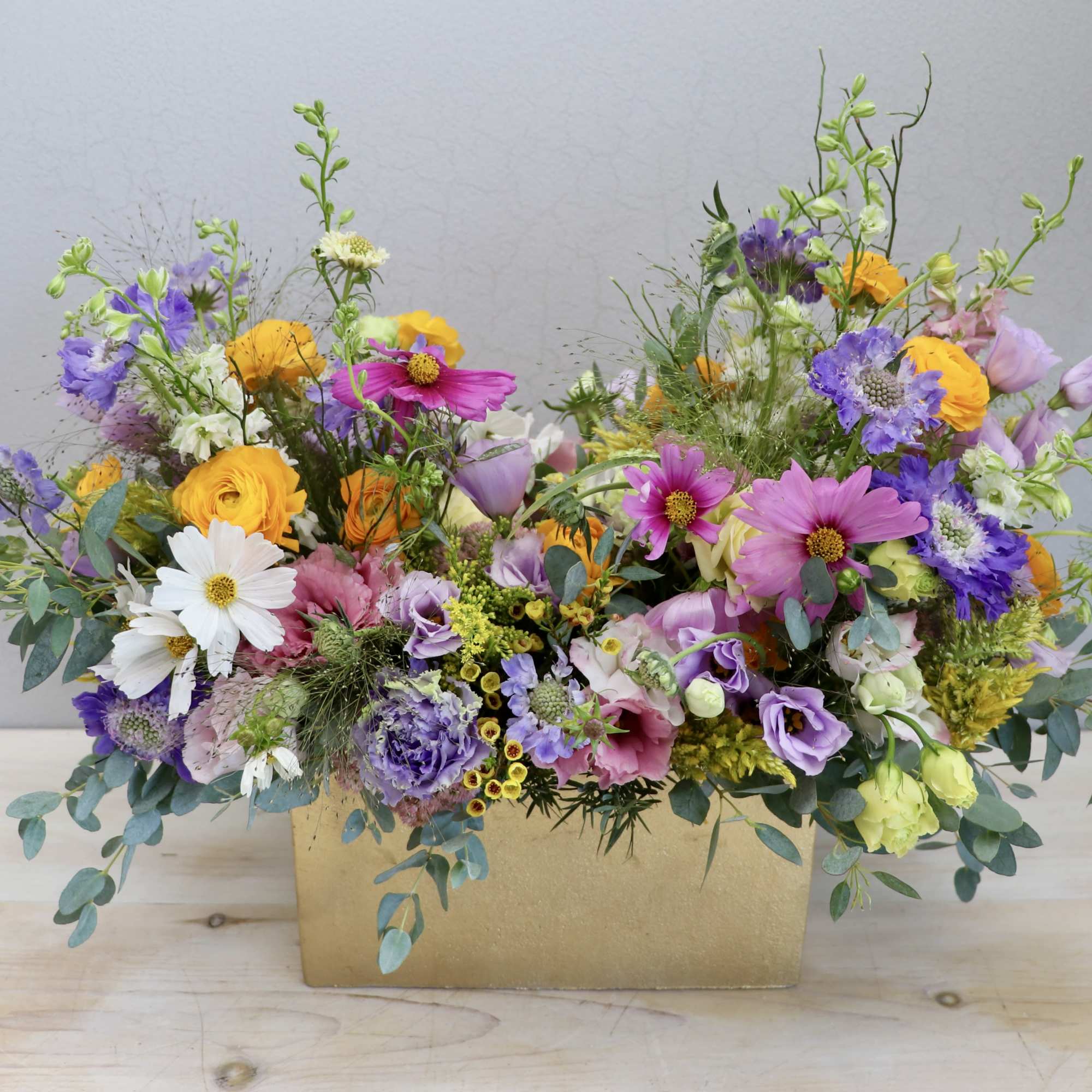 Colorful garden-style arrangement of pink, purple, yellow, and white flowers in a rectangular gold container
