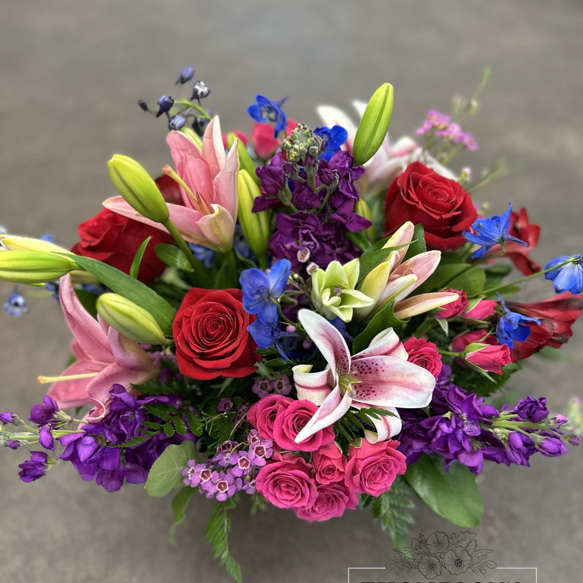 Bright mixed arrangement with pink lilies, red roses, spray roses, and purple and blue accent flowers