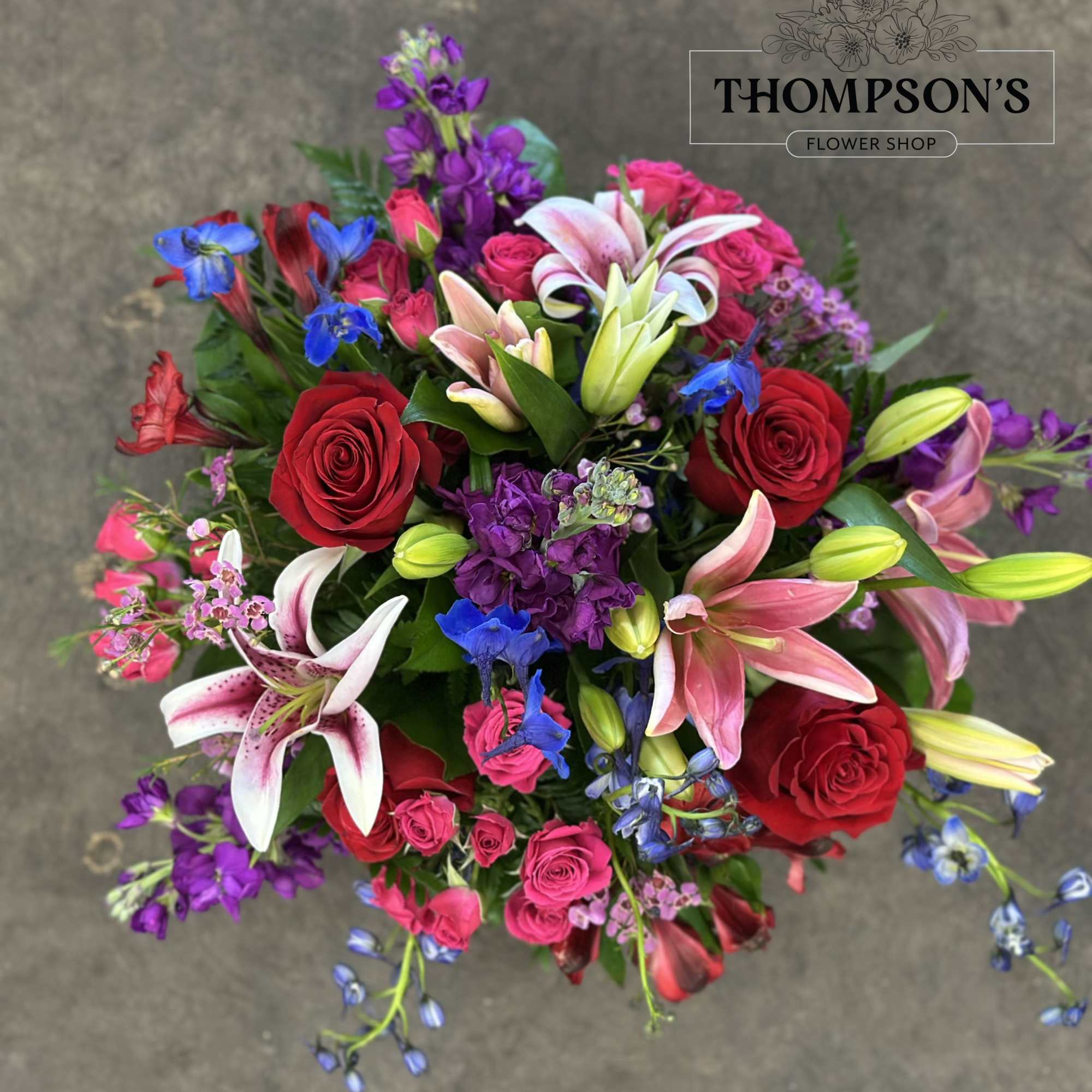 Vibrant mixed bouquet with red roses, pink lilies, and purple and blue flowers arranged in a round design.