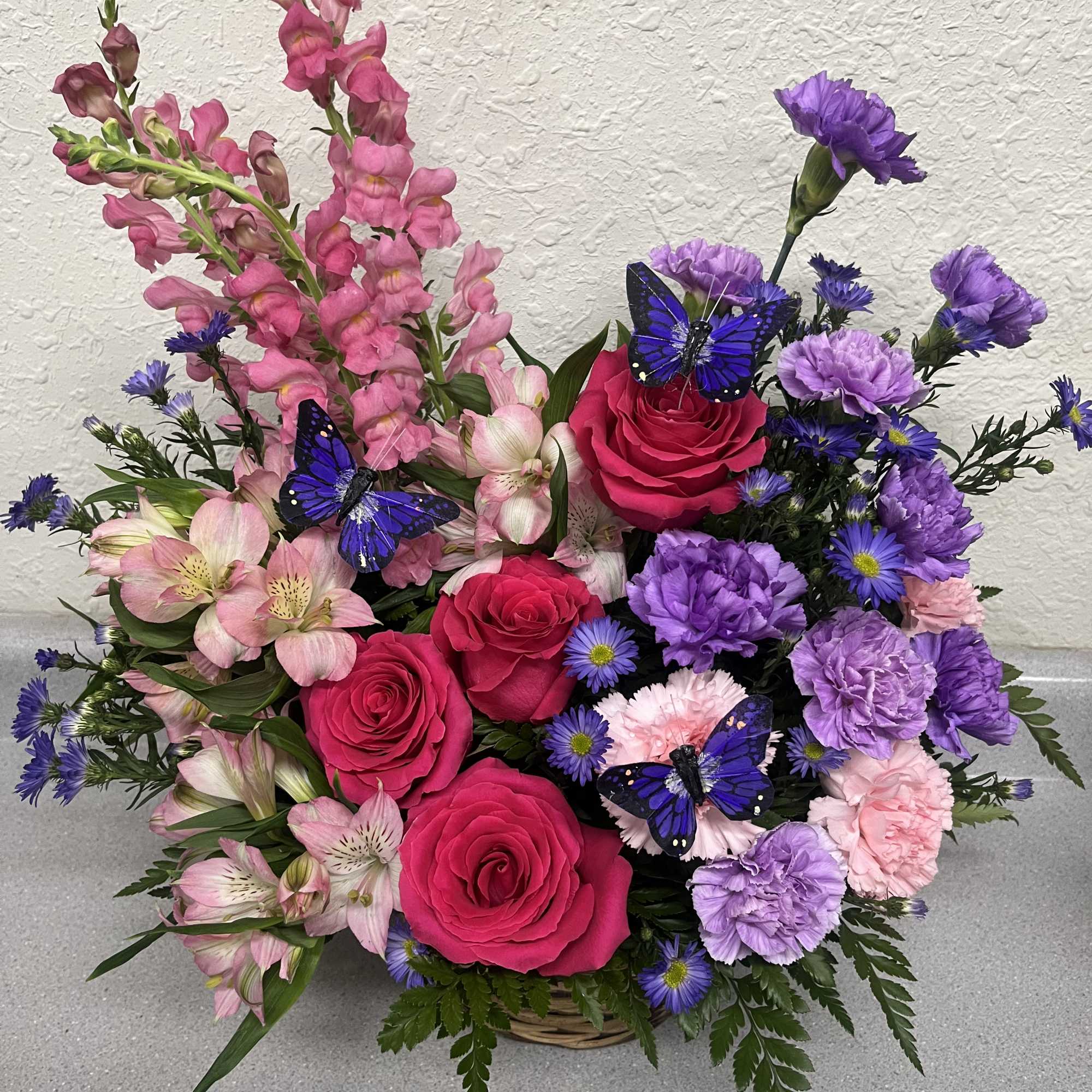 Basket arrangement of pink roses, purple carnations, and pink snapdragons with butterfly picks