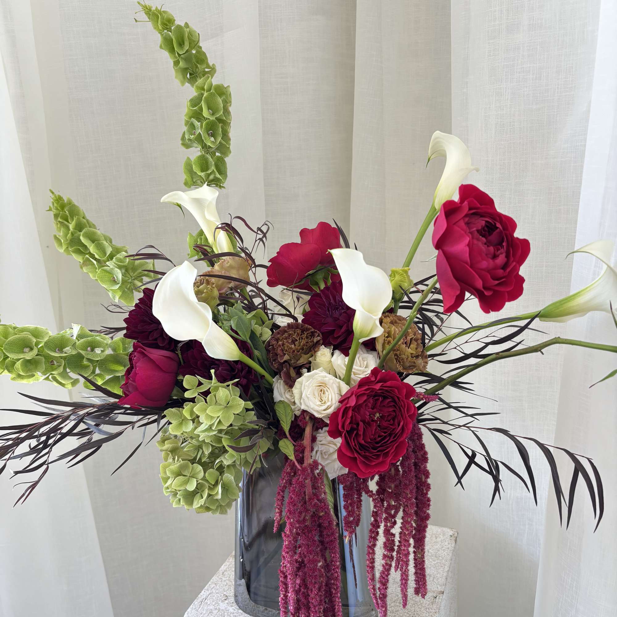 Tall floral arrangement with red roses, white calla lilies, and green hanging blooms in a glass vase