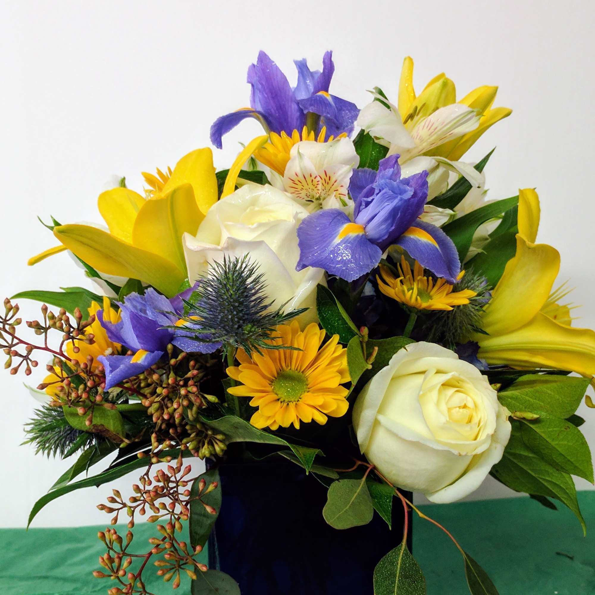 Mixed bouquet of yellow lilies and daisies, purple irises, and white roses in a dark blue glass vase