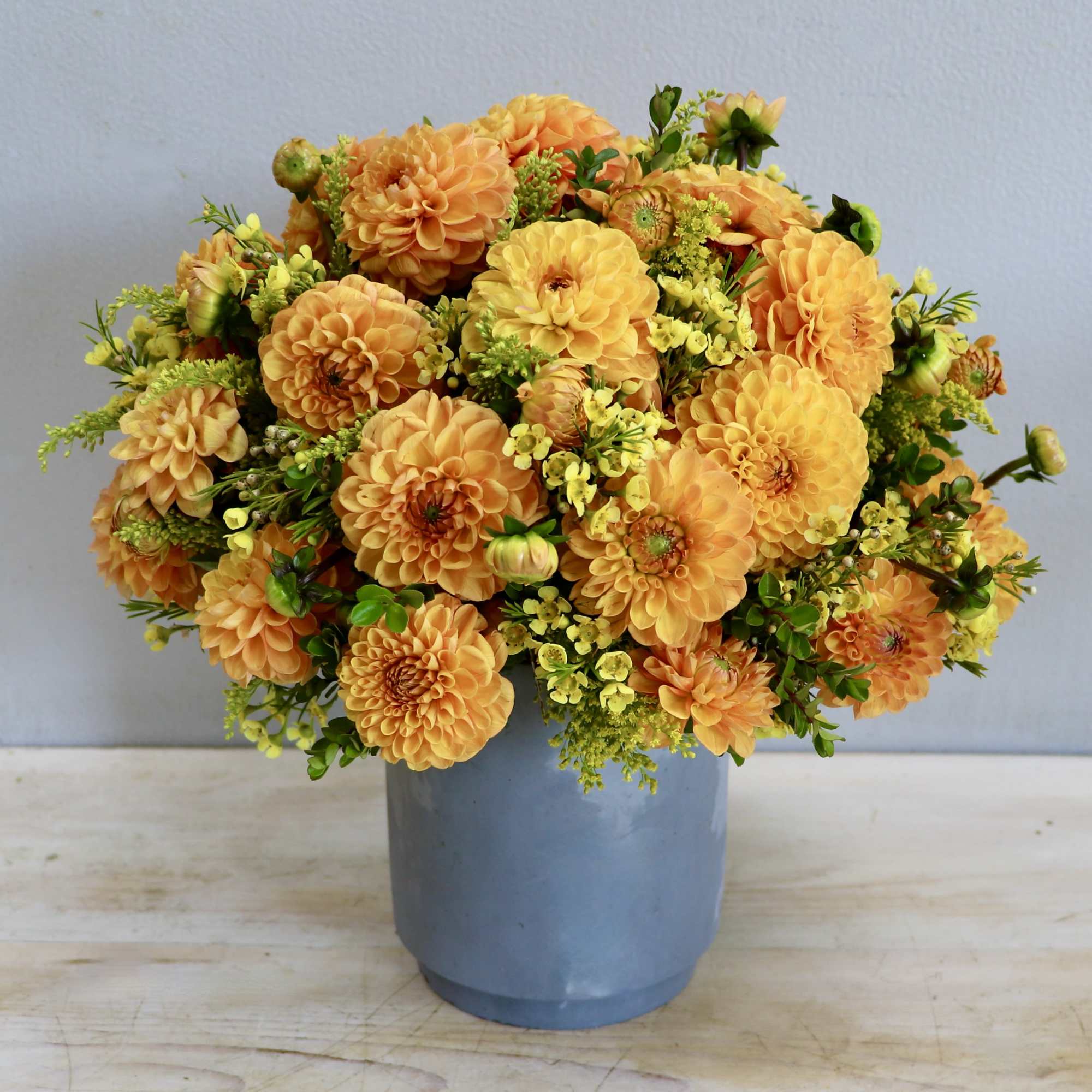 Compact arrangement of orange dahlias and yellow blooms in a matte blue-gray vase
