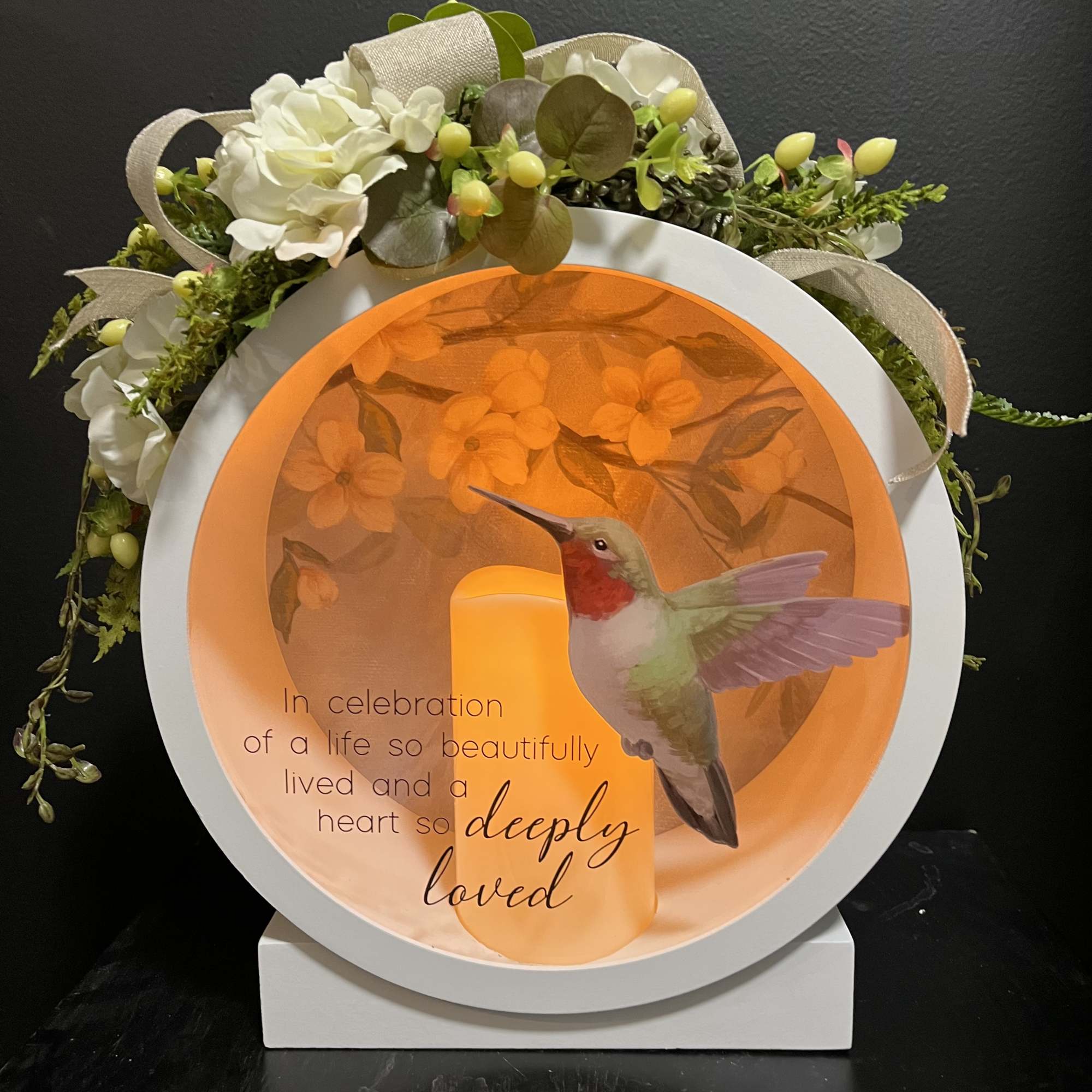 Circular memorial display with white flowers, ribbons, and a hummingbird candle