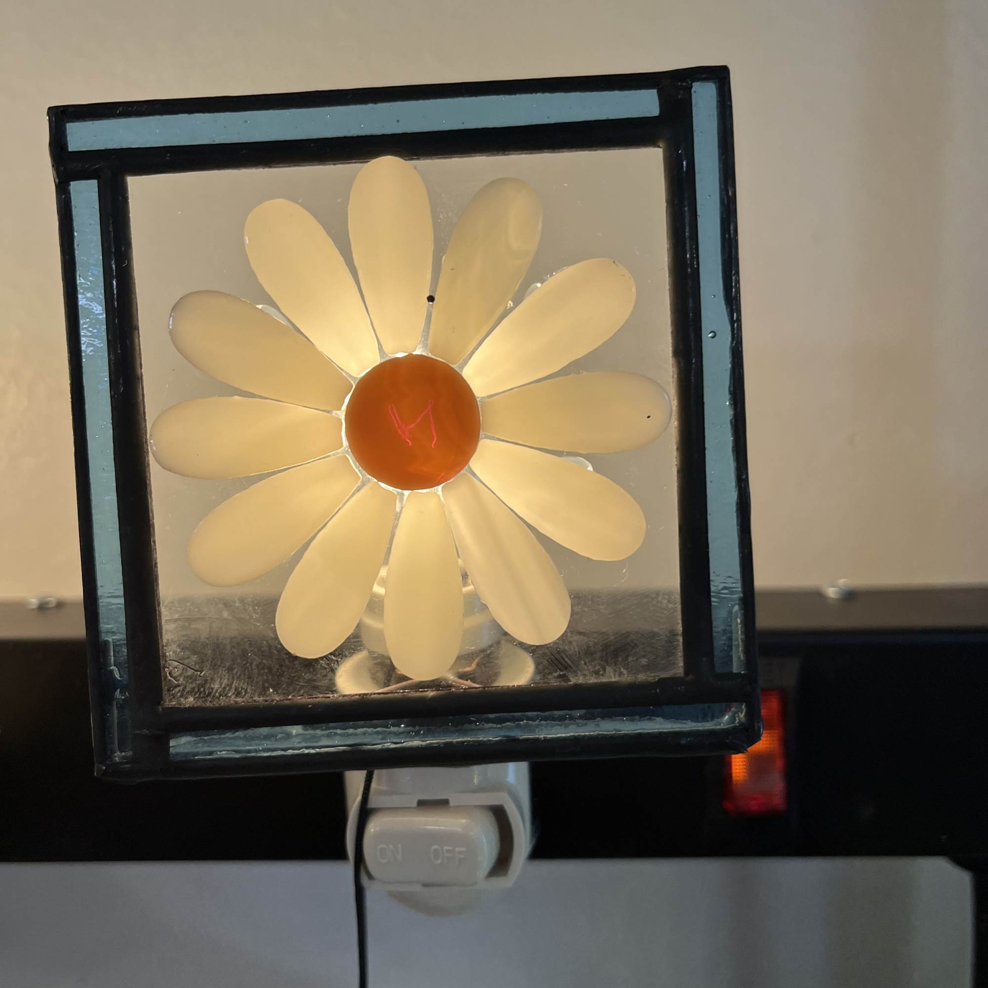 Lit daisy-shaped lamp with white petals and an orange center in a square frame