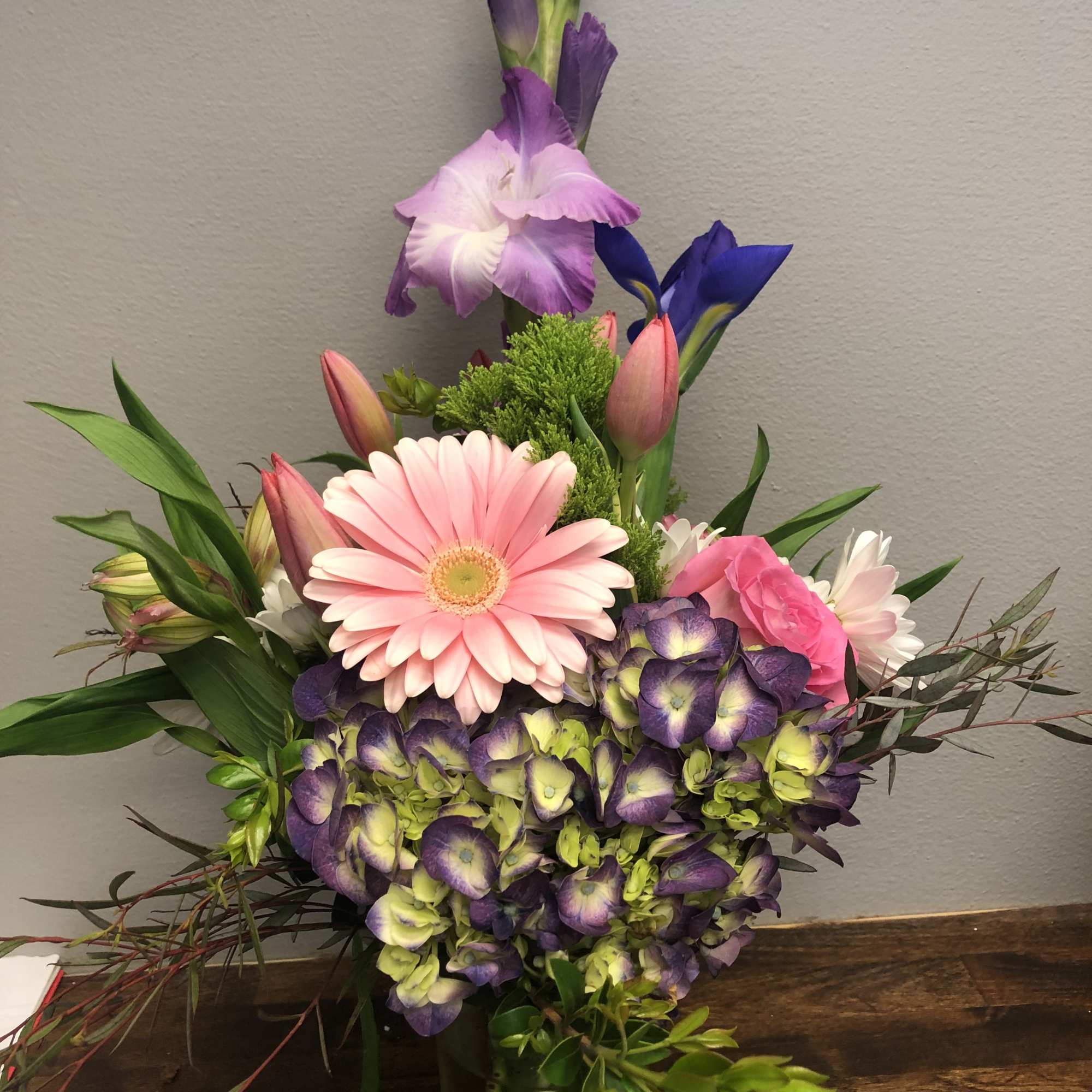 Mixed bouquet with pink gerbera daisies, purple blooms, and a tall iris in a glass vase