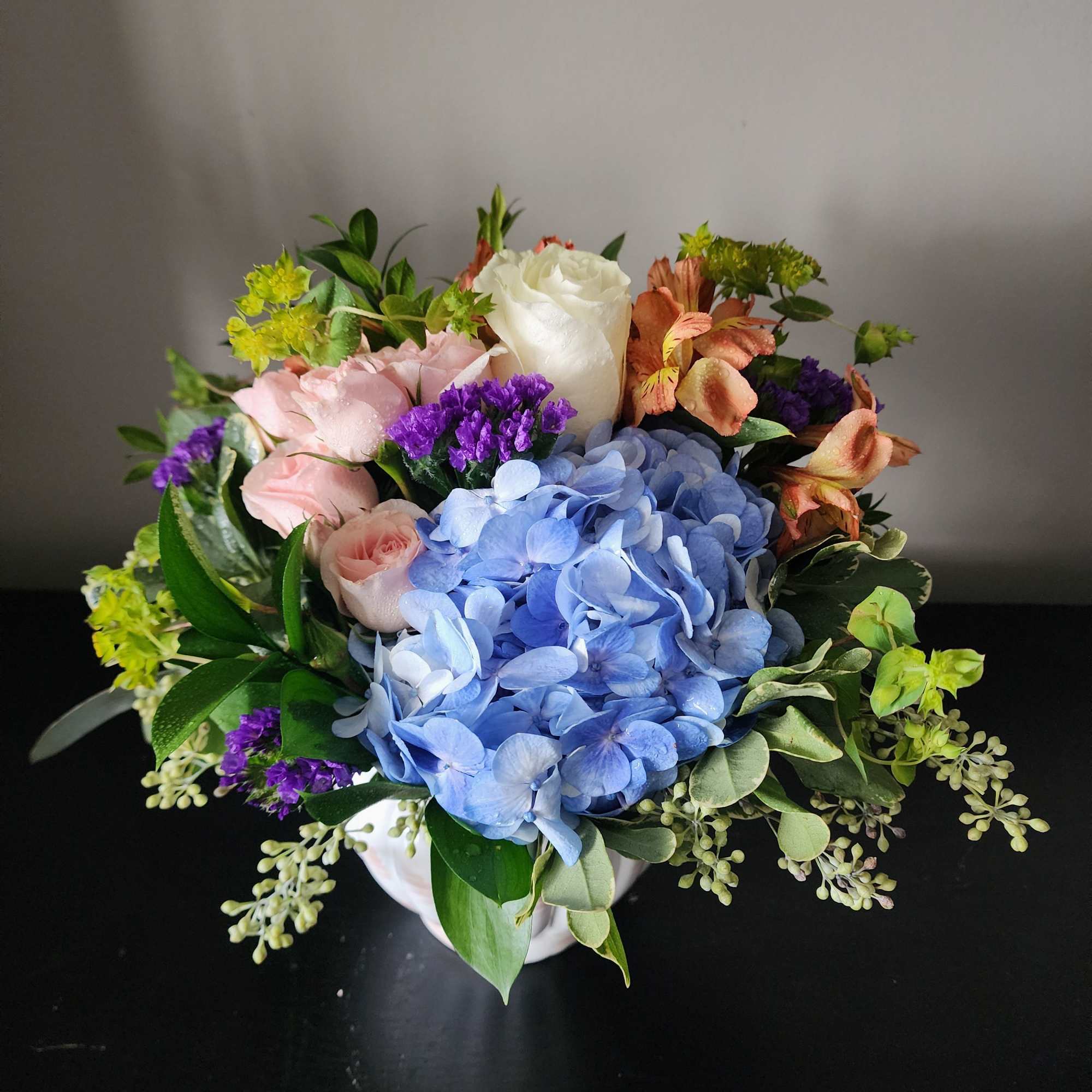 Mixed bouquet with blue hydrangea, roses, and purple accents in a white vase