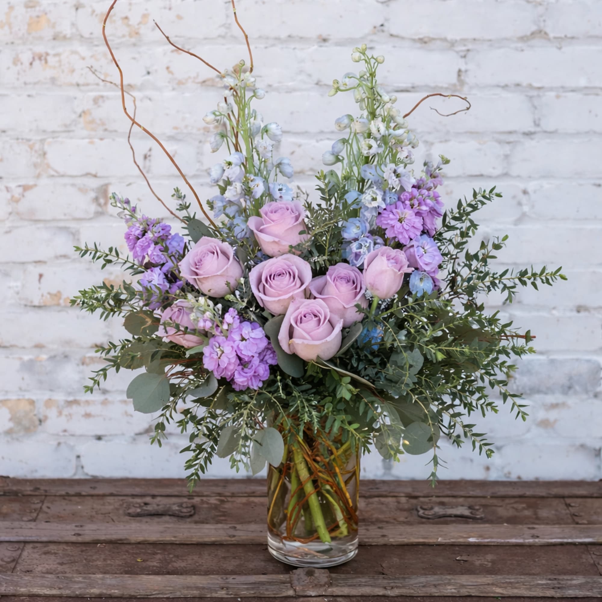 Lavender roses and purple flowers arranged in a clear glass vase.