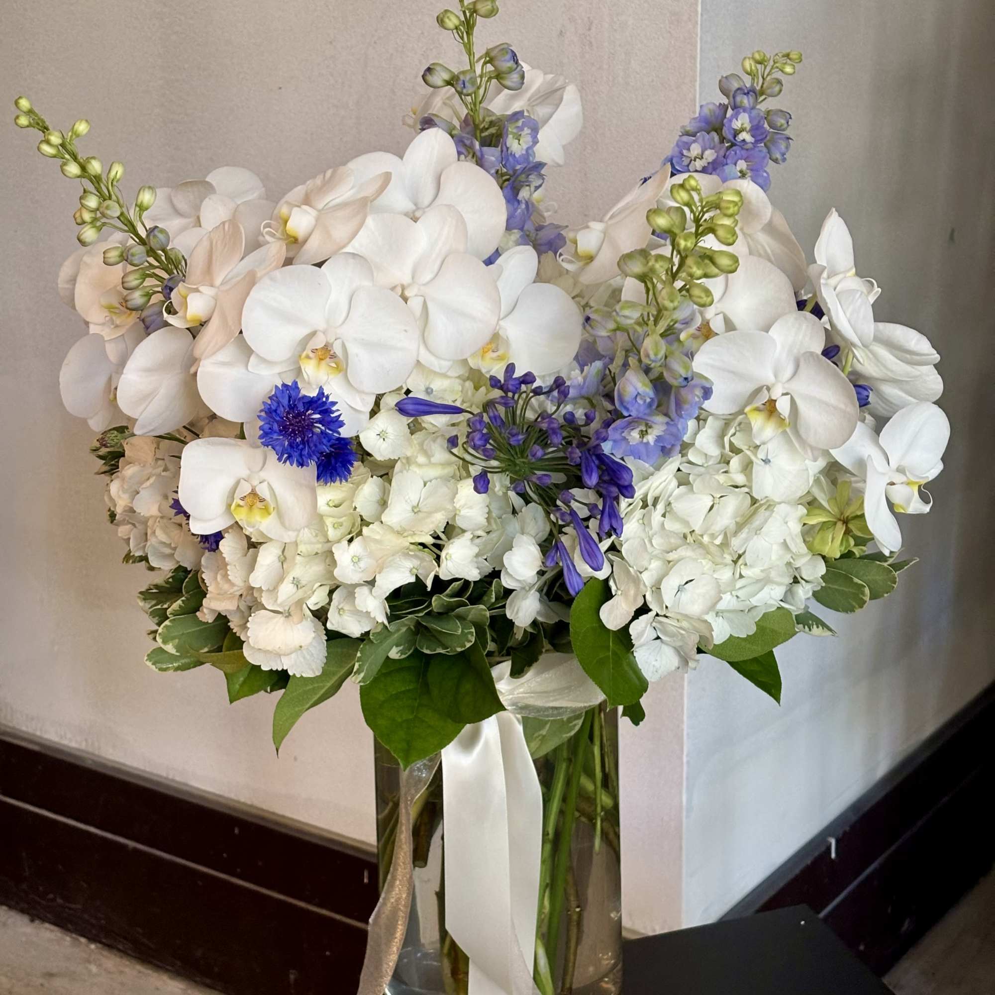 White orchids and blue flowers arranged in a tall glass vase with ribbon