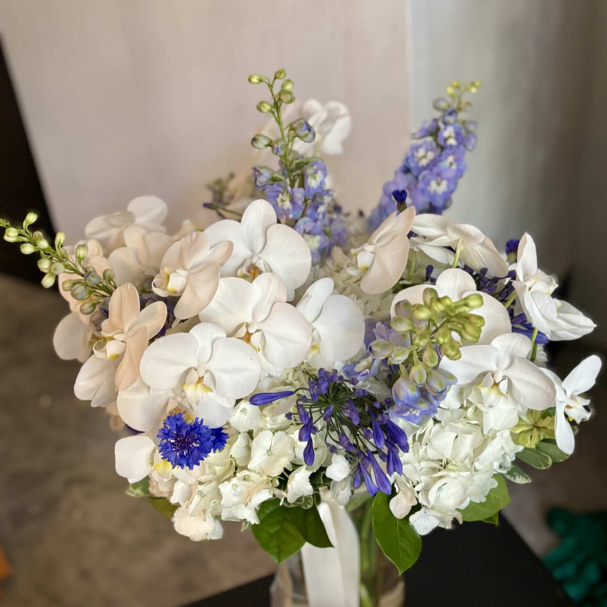 White orchids and blue flowers arranged in a glass vase with ribbon