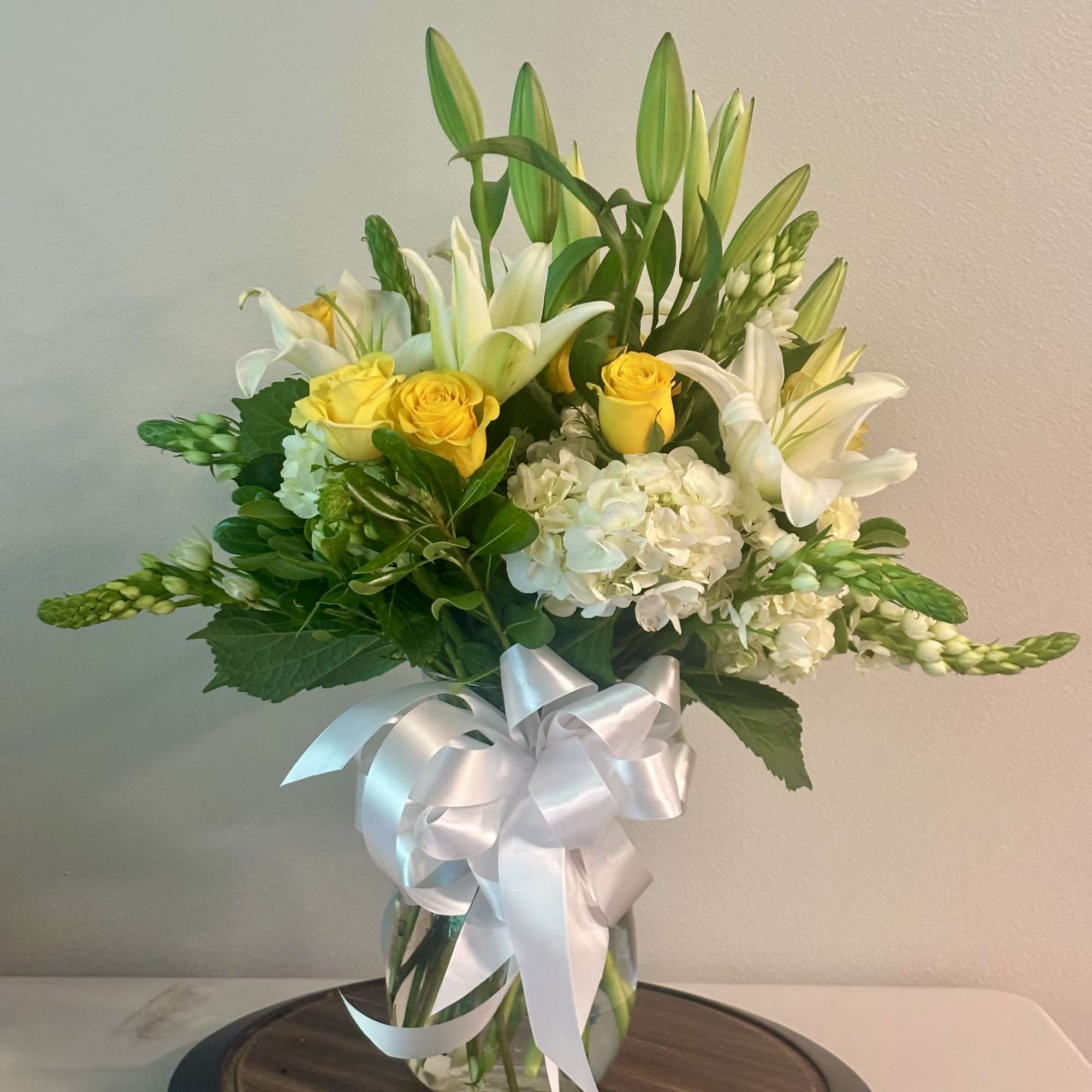 Yellow roses and white lilies in a glass vase with a white ribbon