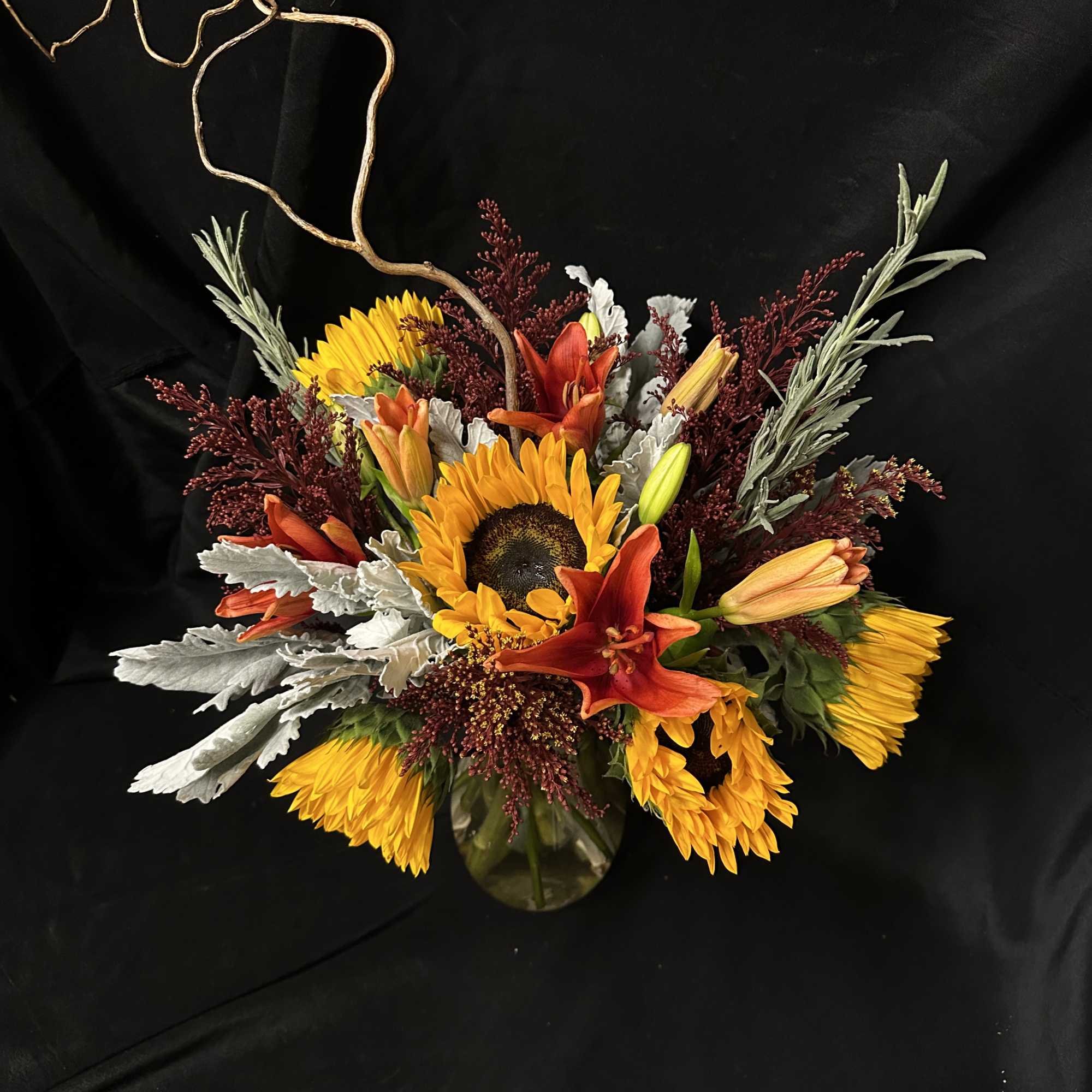 Sunflowers and orange lilies arranged in a glass vase with curly branches