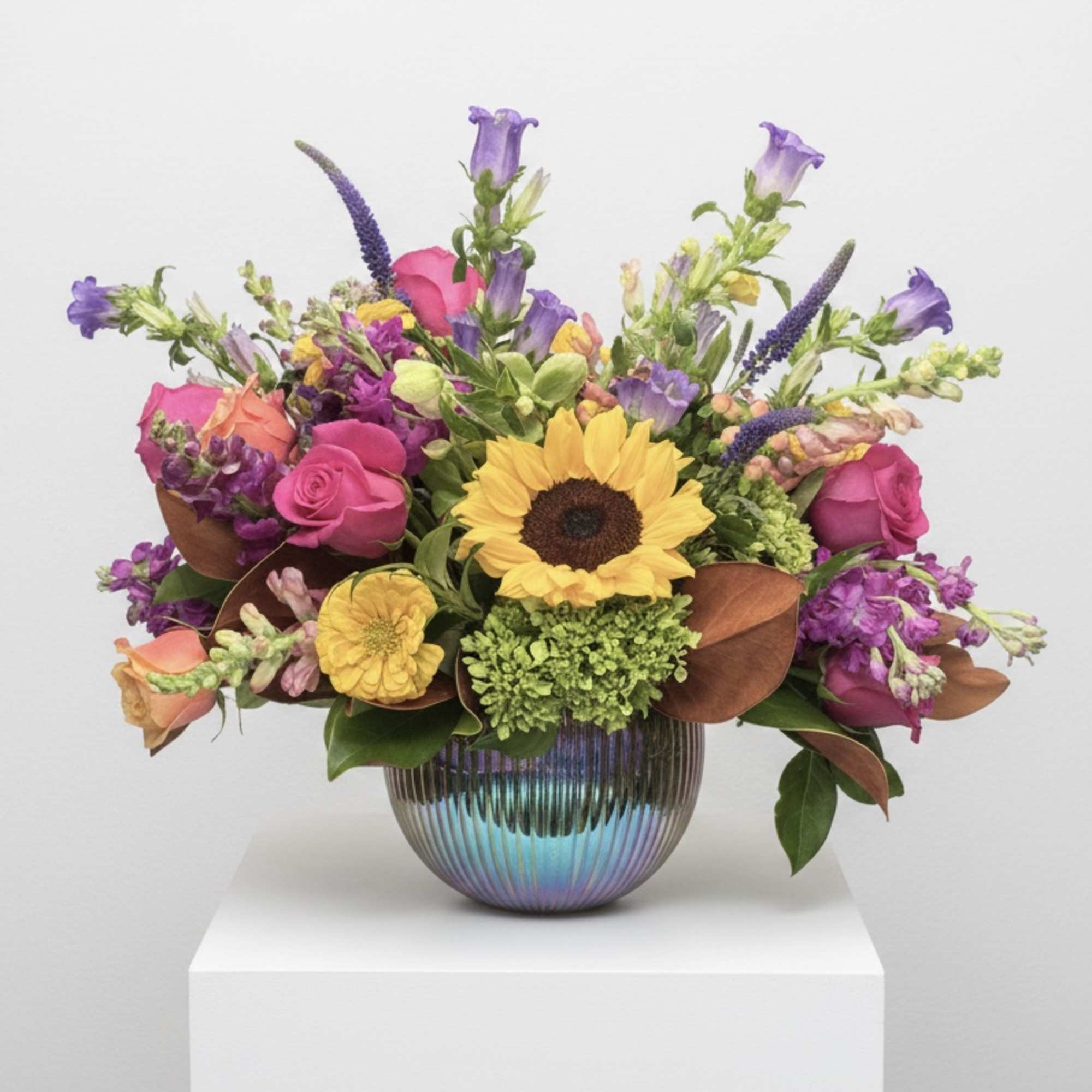Mixed bouquet with a sunflower, pink roses, and purple blooms in a ribbed iridescent vase