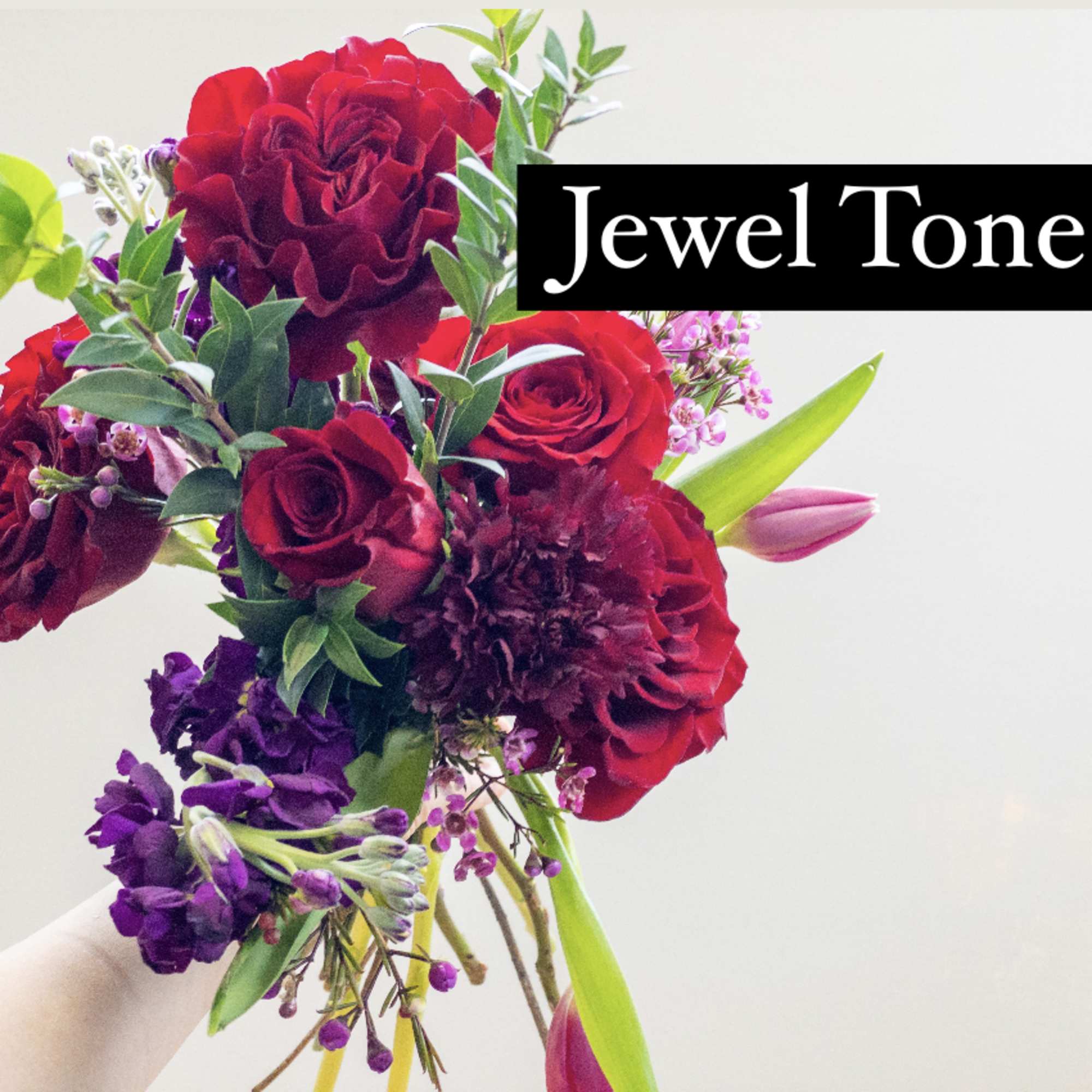 Handheld bouquet of red roses and jewel-toned flowers against a light background