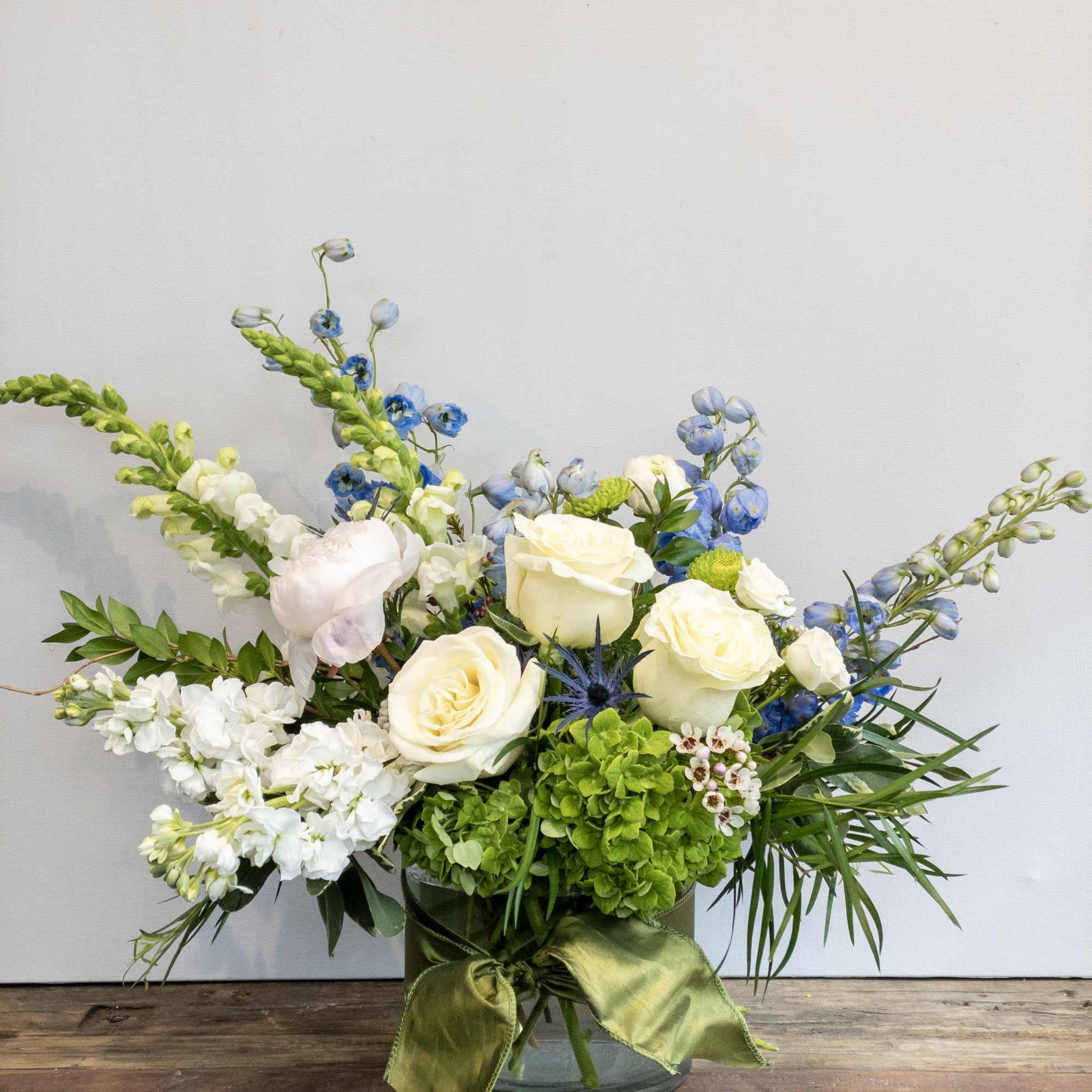 Low arrangement of white roses, blue delphinium, and green hydrangea in a glass vase with ribbon