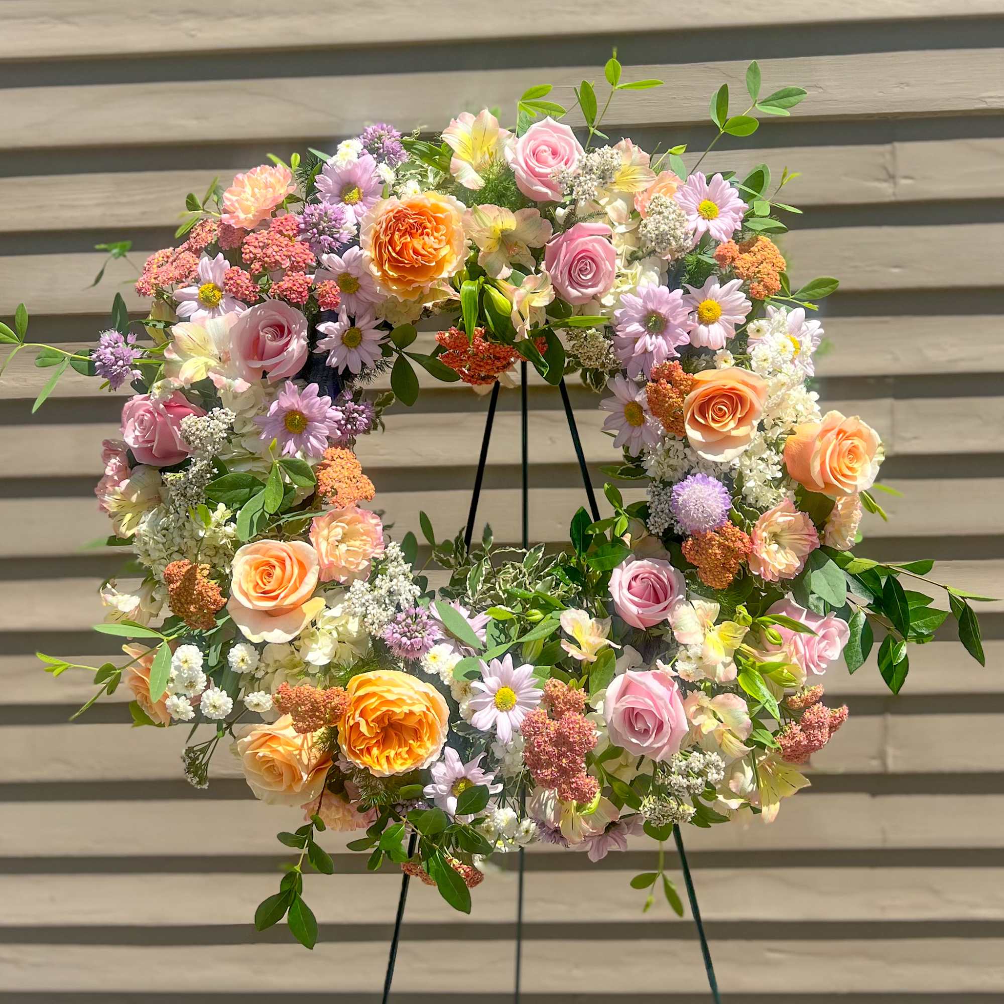 Pastel wreath of roses, daisies, and mixed blooms on a metal easel against a neutral wall.