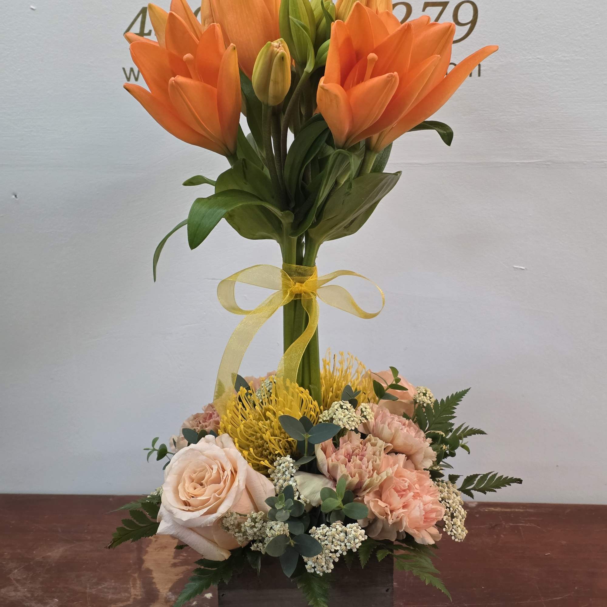 Orange lilies above a low arrangement of peach roses and yellow blooms in a wooden box