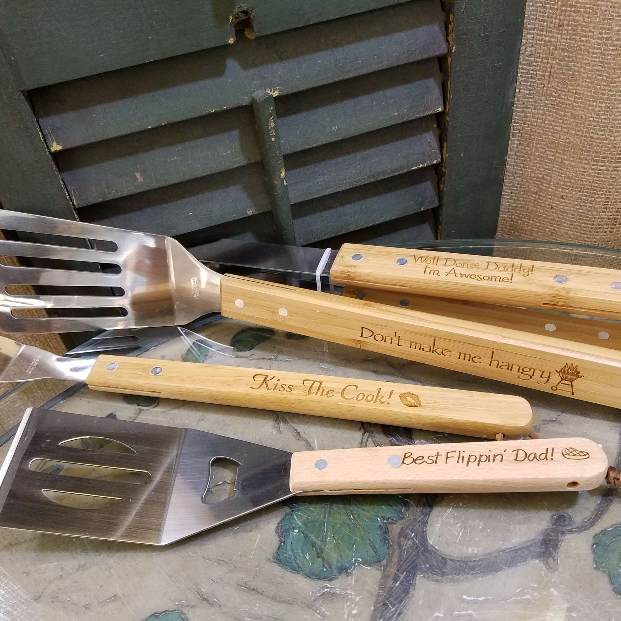 Four engraved grilling spatulas with wooden handles on a glass table.