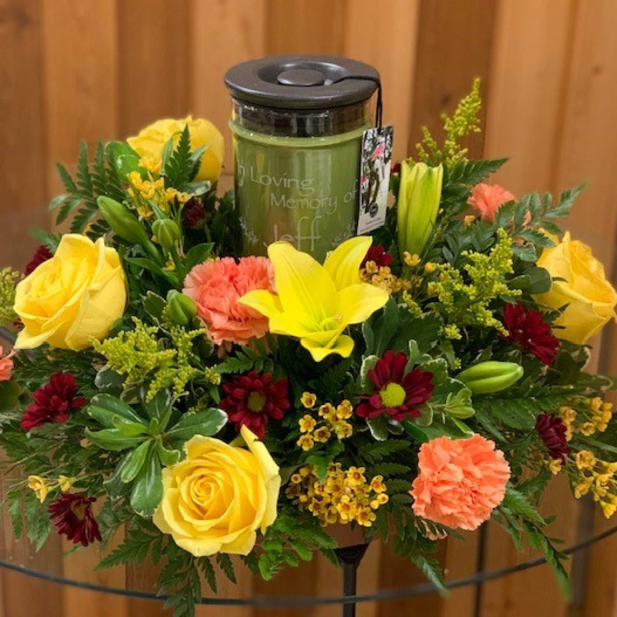 Yellow roses and lilies surround a memorial candle in a floral arrangement.