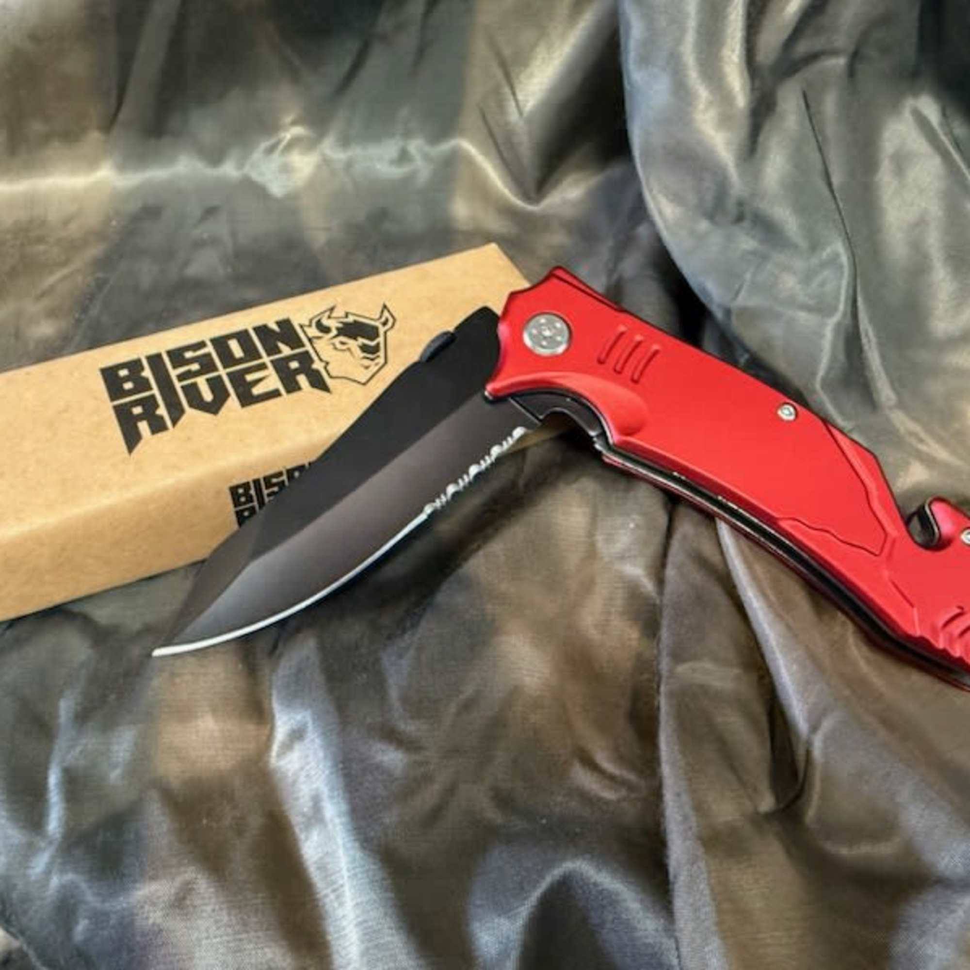 Red folding knife beside a branded cardboard box on dark fabric