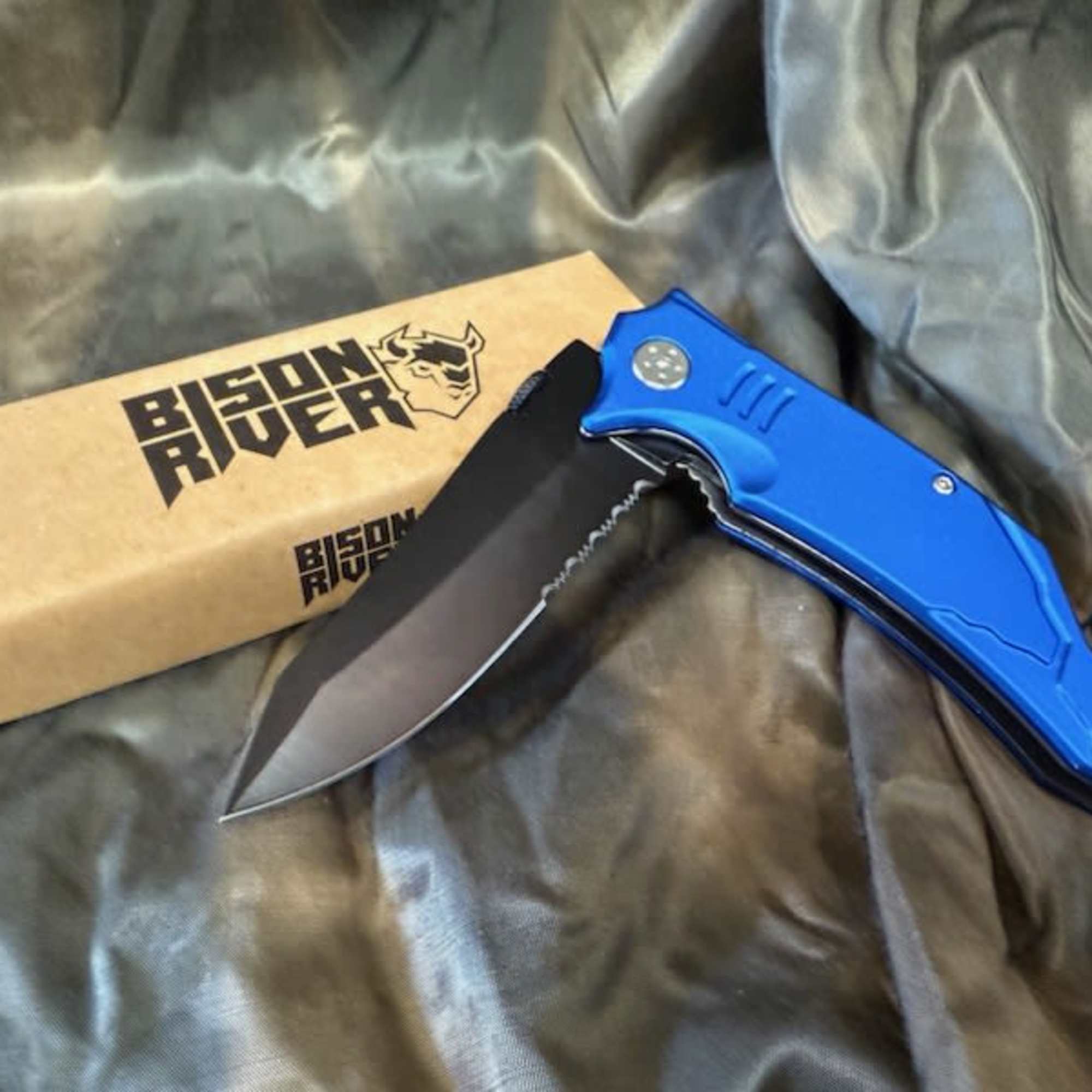 Blue folding knife beside a branded cardboard box on dark fabric