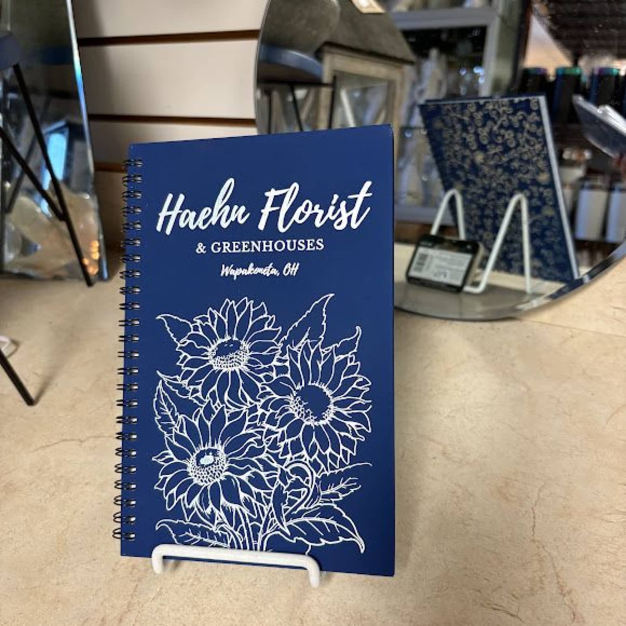 Blue spiral notebook with white sunflower line art on a display stand