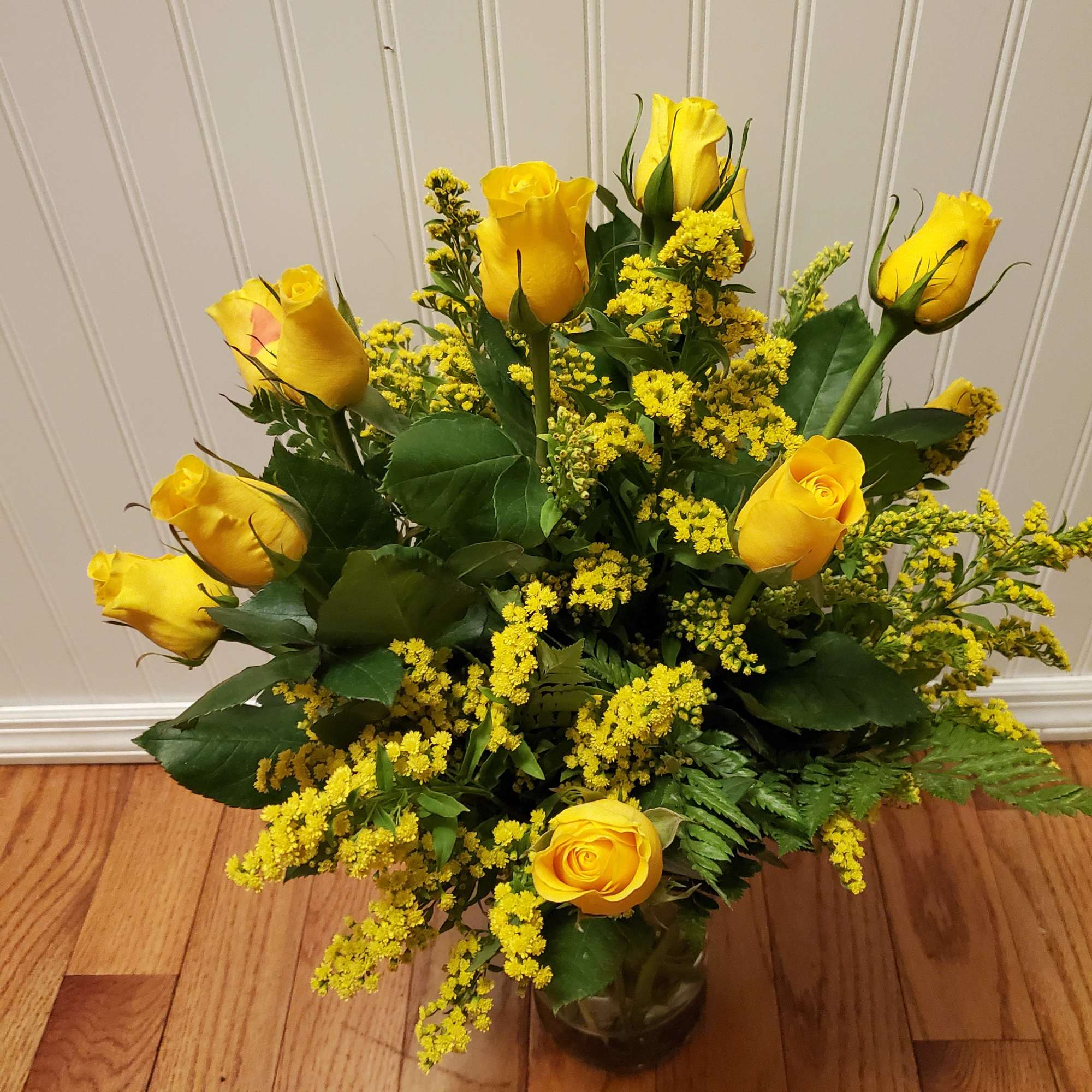 Vase arrangement of yellow roses with yellow filler flowers in a clear glass vase on a wooden floor
