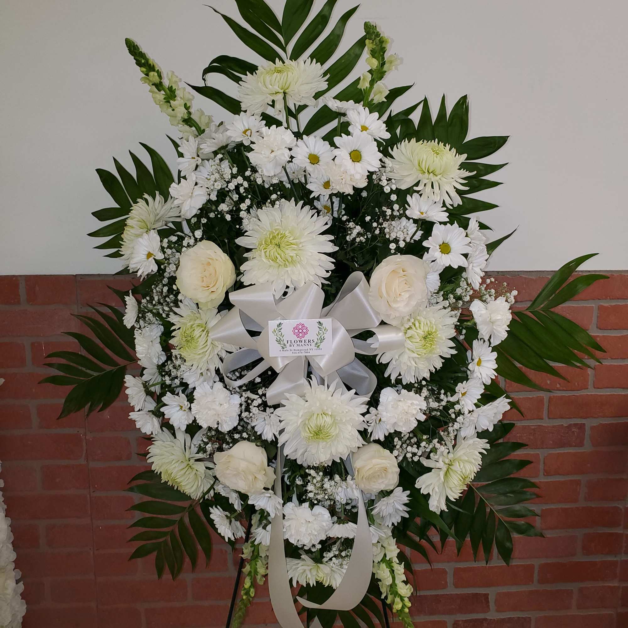 Standing white funeral spray of roses, daisies, and mums with a white ribbon on an easel.
