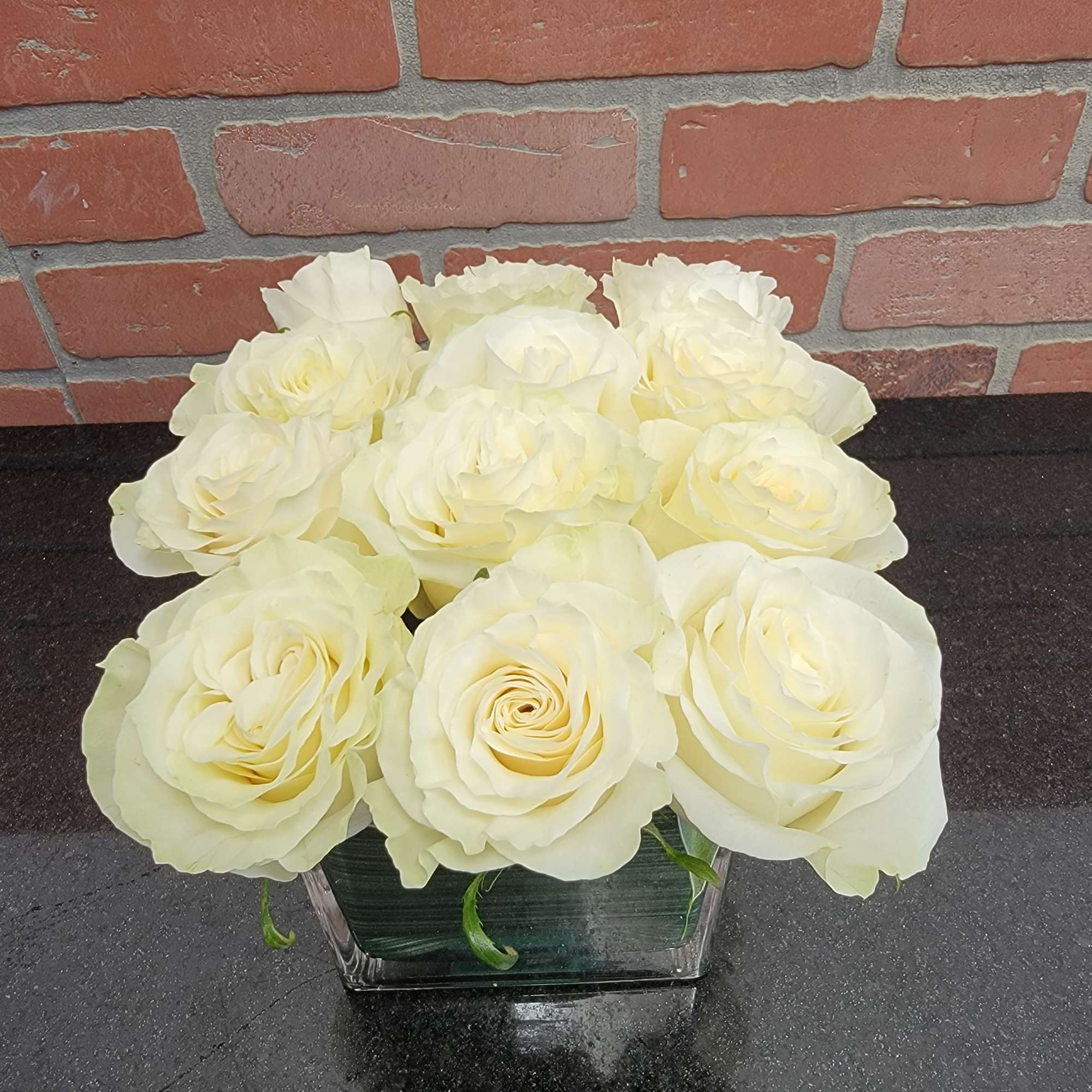 Low arrangement of pale cream roses in a square clear glass vase on a dark countertop
