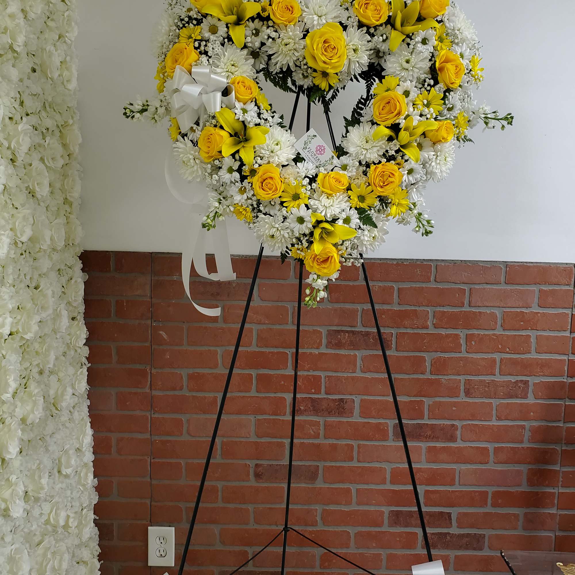 Heart-shaped yellow and white standing wreath on a black easel with white ribbon bows
