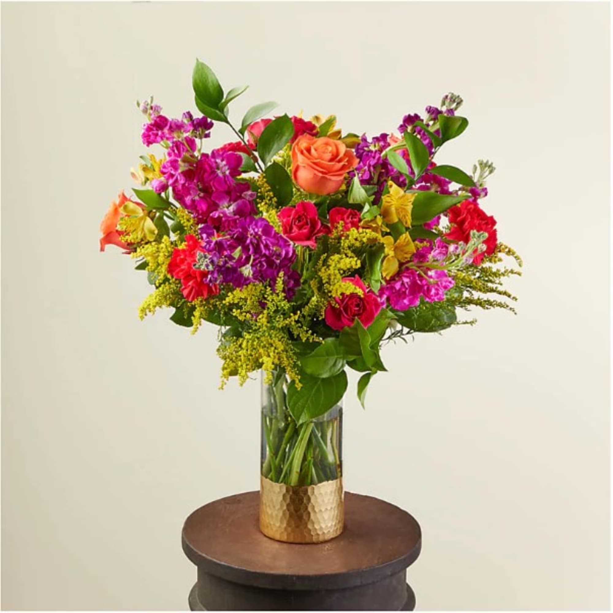 Tall mixed bouquet of orange roses, pink stock, red spray roses and yellow flowers in a clear vase with gold base.