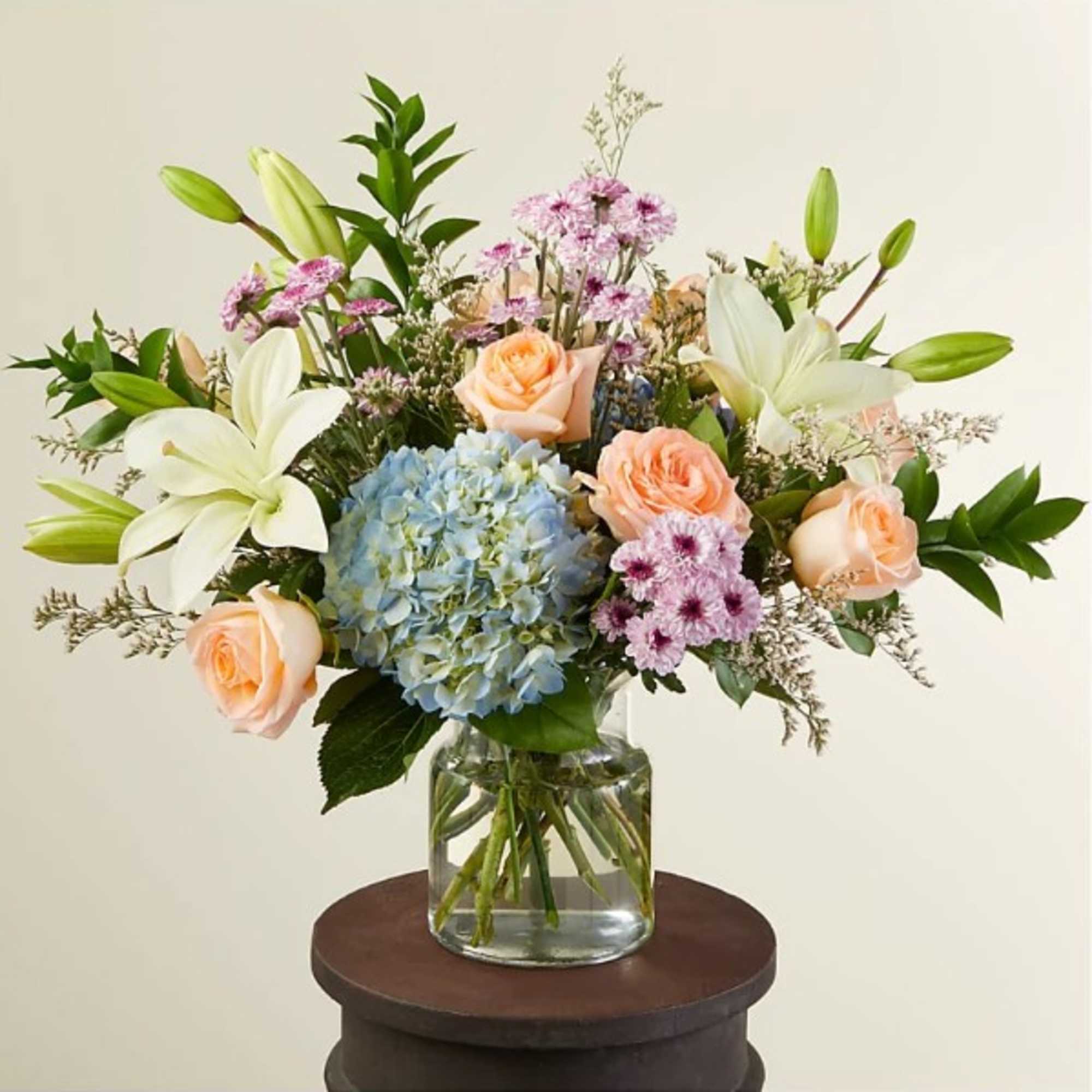 Mixed bouquet of peach roses, white lilies, blue hydrangea and lavender mums in a clear glass vase