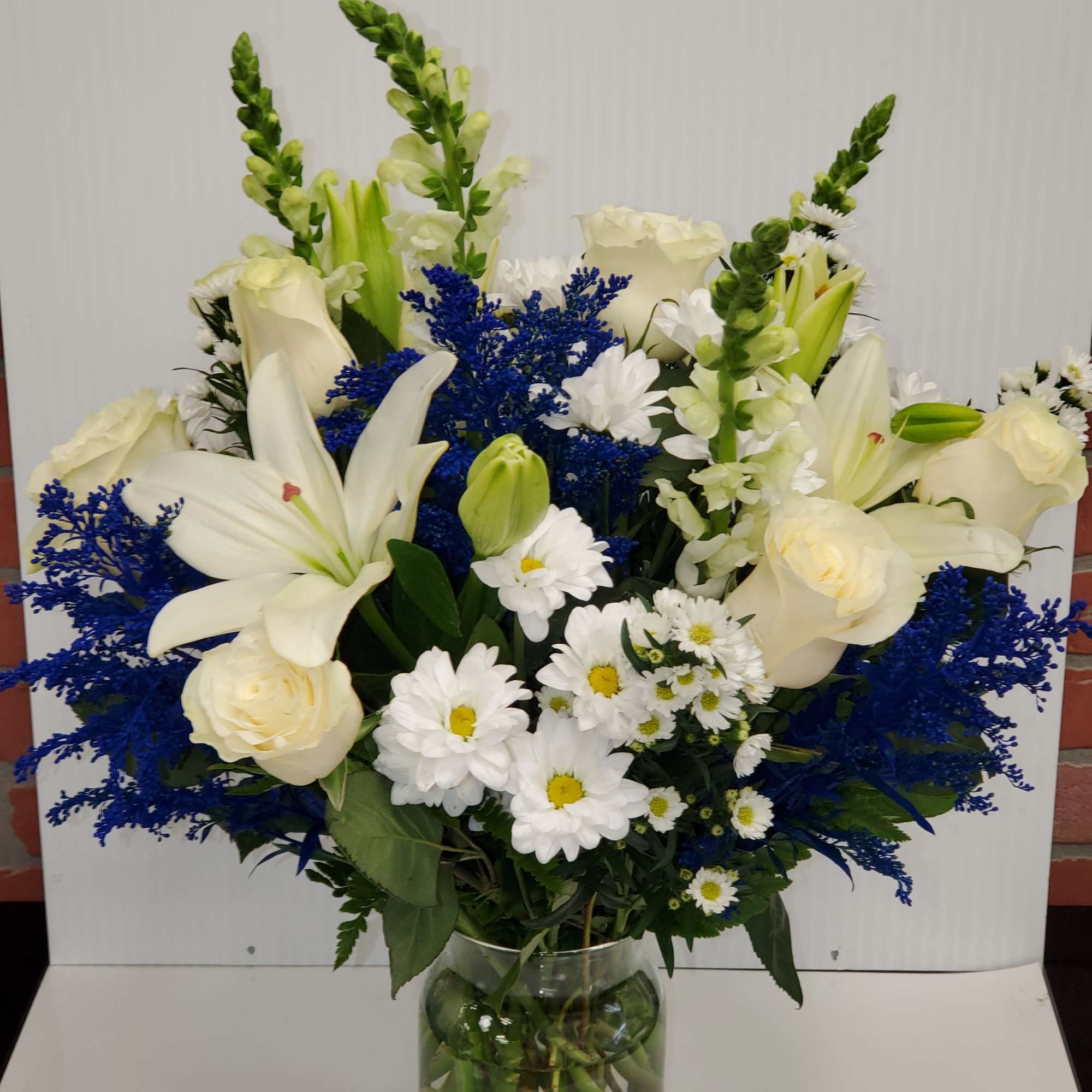 Tall vase arrangement of white lilies, roses, and daisies with bright blue filler flowers