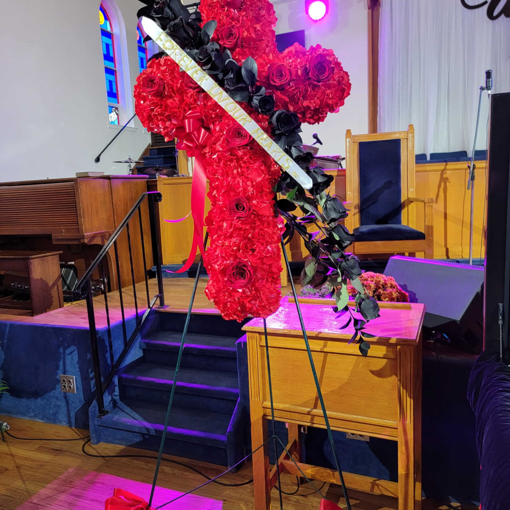 Standing red floral cross with roses and black accents on a metal easel in a church.