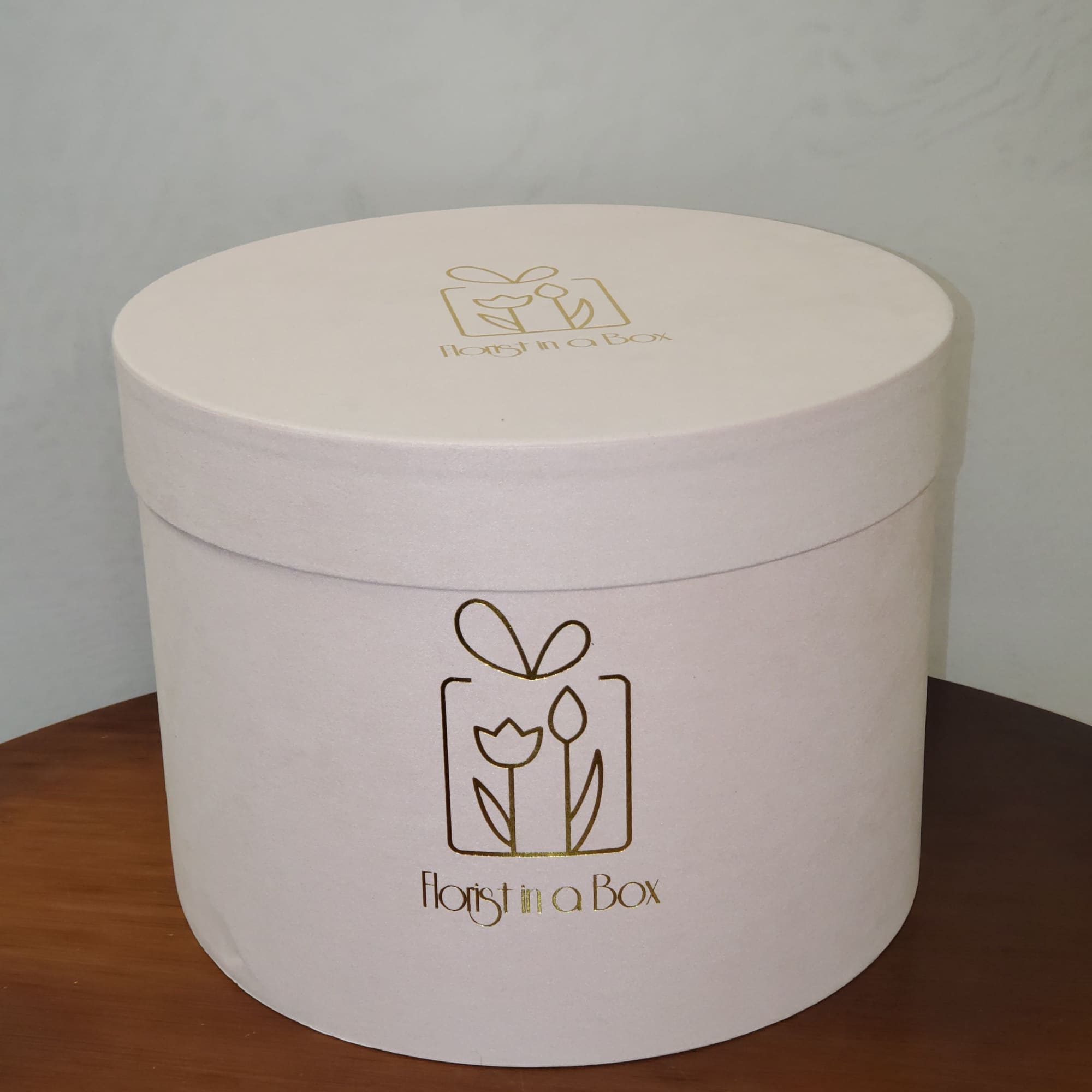 Round off-white hatbox-style gift box with gold floral logo on a wooden surface