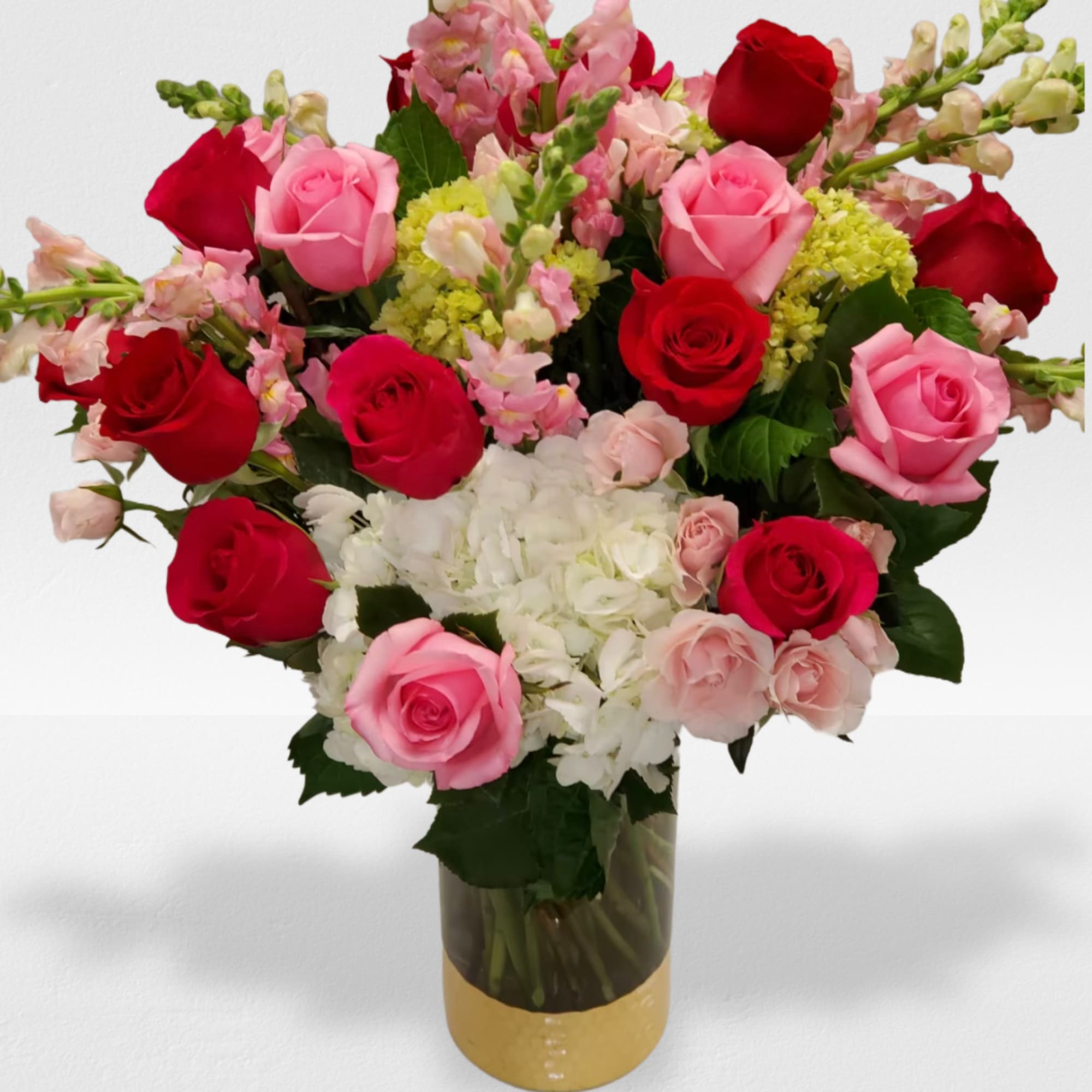 Arrangement of red and pink roses with white hydrangeas and pink snapdragons in a glass vase with a gold base