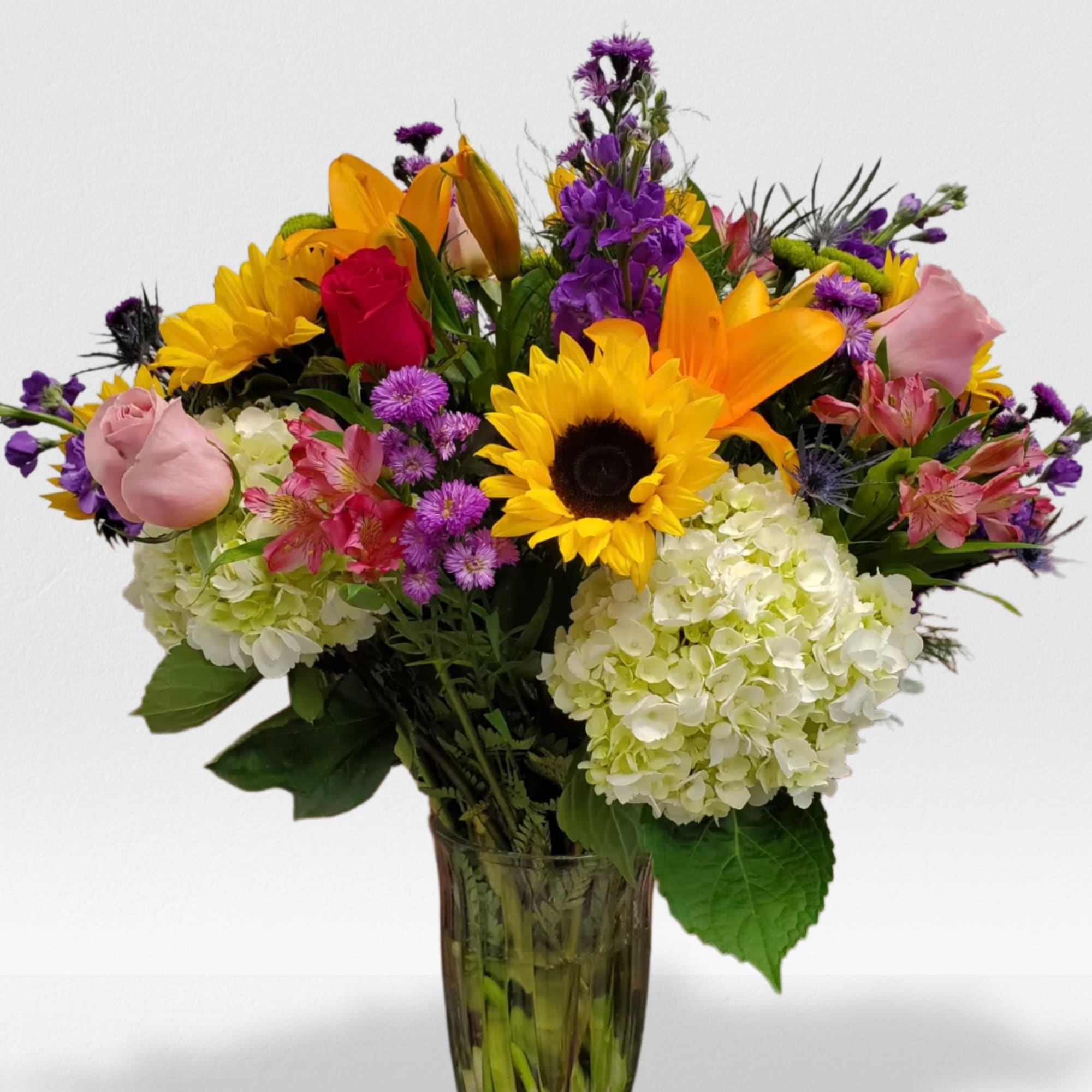 Tall mixed bouquet with sunflowers, roses, lilies, hydrangeas and purple flowers in a clear glass vase