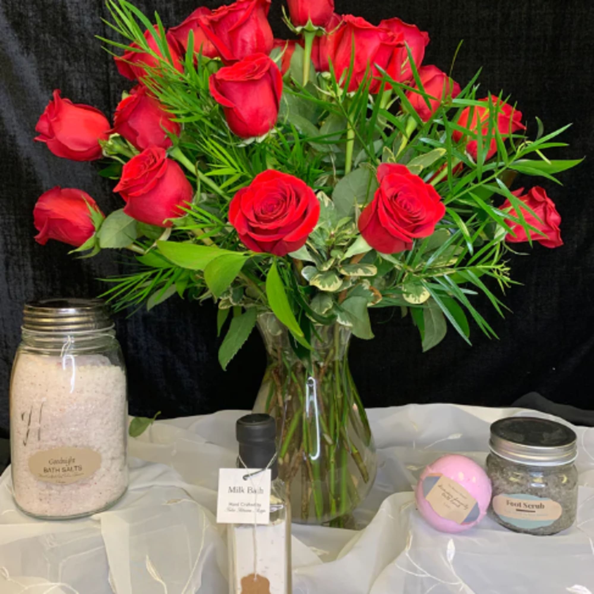Red roses arranged in a clear glass vase with assorted bath gift items