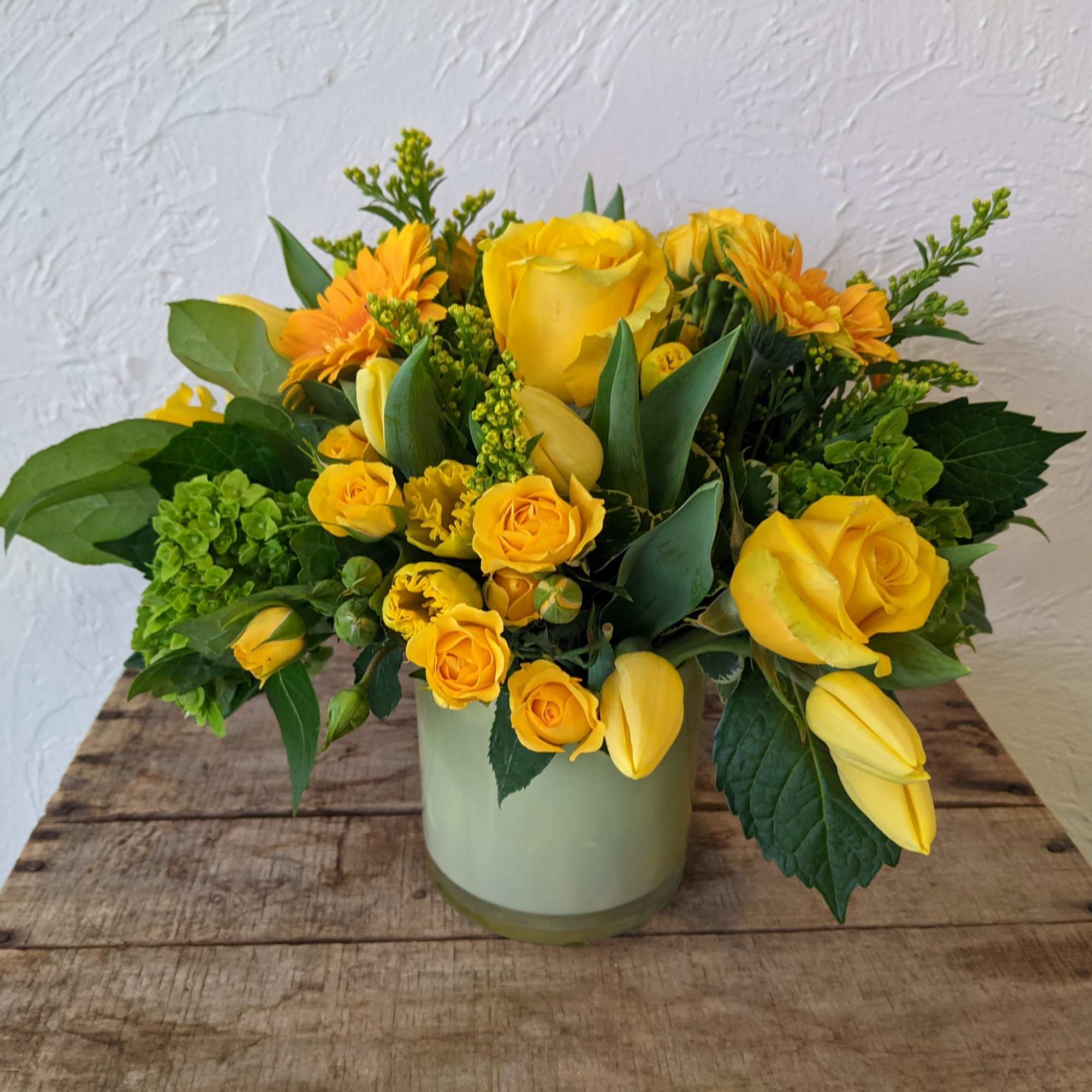 Low yellow rose, tulip, and gerbera daisy arrangement in a pale green glass cylinder vase