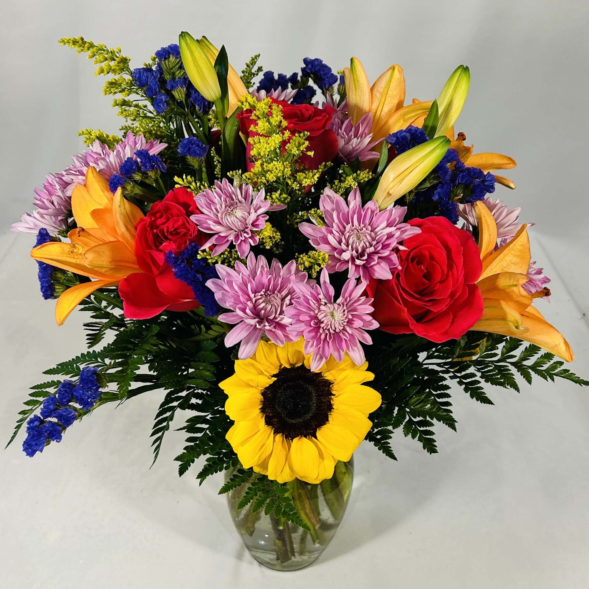 Bright mixed bouquet with sunflower, red roses, orange lilies, and purple mums in a clear glass vase