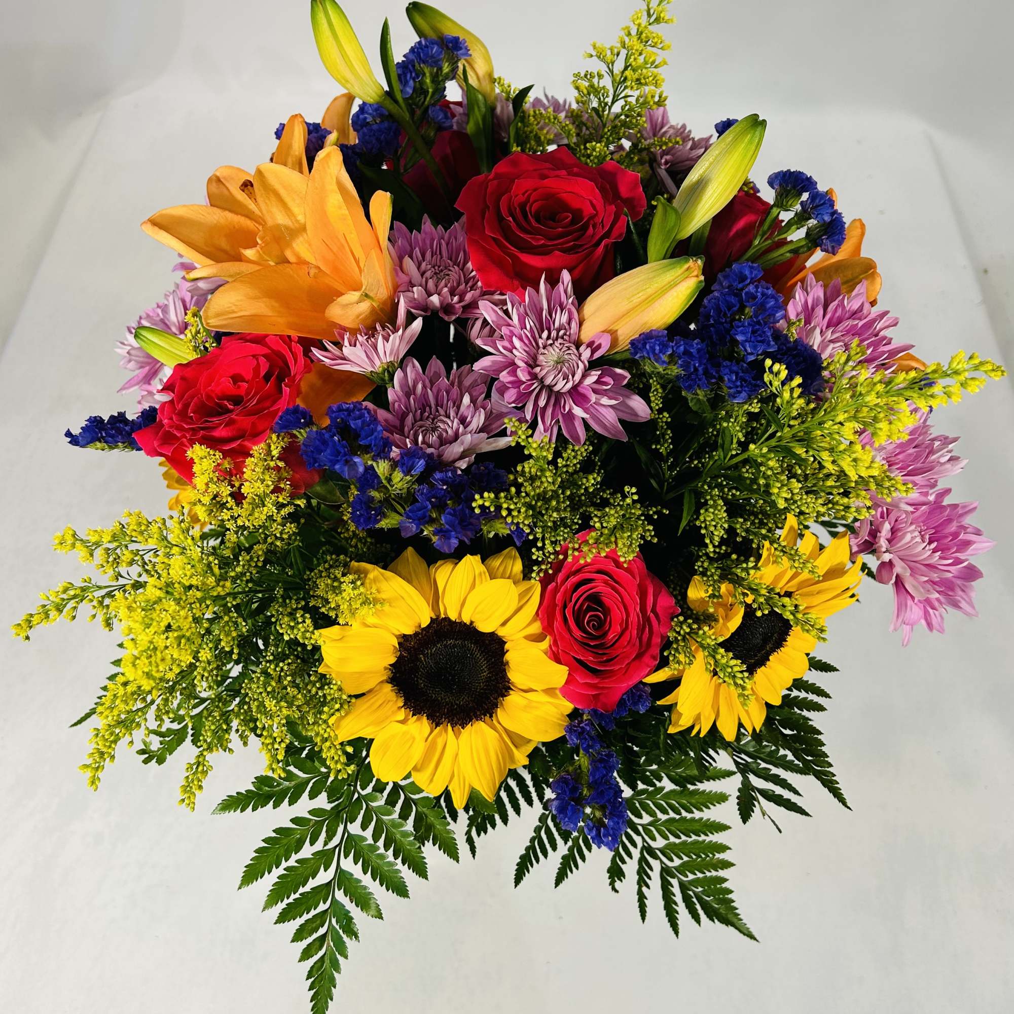 Bright mixed bouquet with sunflowers, red roses, orange lilies, purple mums, and blue accents in a compact design